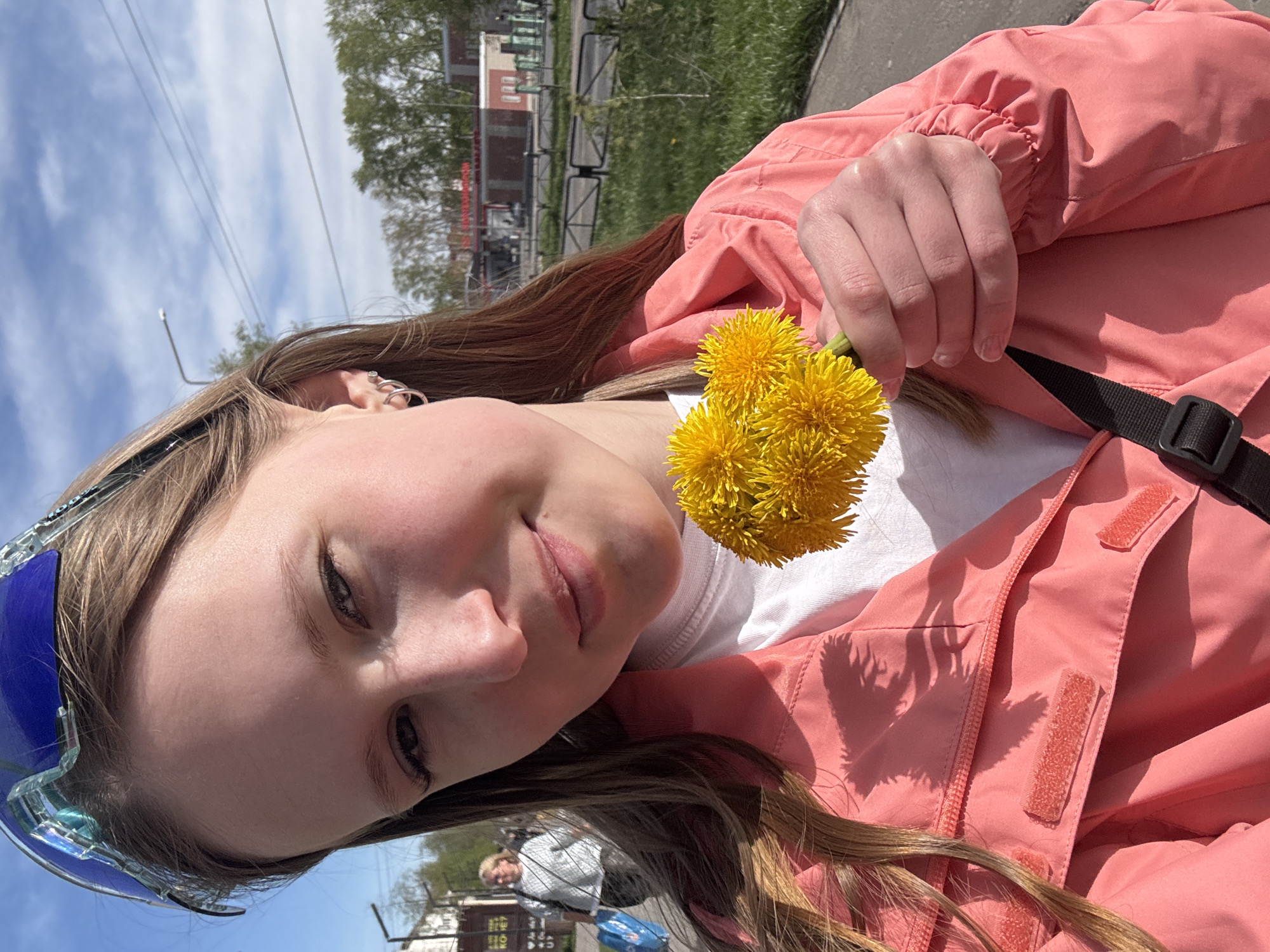 A young person is holding three yellow dandelions. The person is wearing a white shirt and a coral-colored jacket with a black strap. They also have blue sunglasses on their head and multiple earrings. The background shows a cityscape with buildings, grass, and a cloudy sky. The location is identified as Chaikovsky, Russia. Other people are visible in the far background.