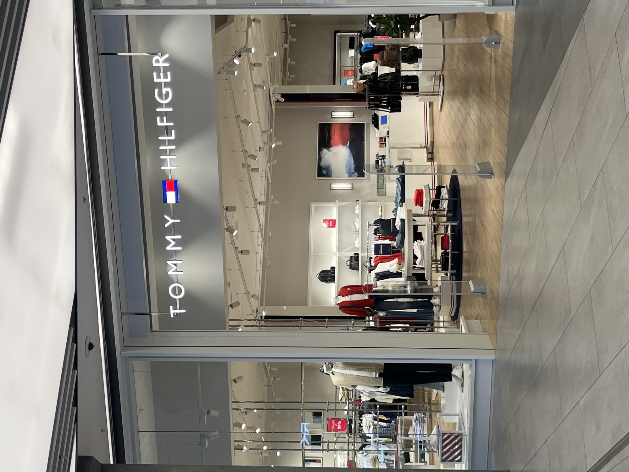 The image shows the exterior of a Tommy Hilfiger store located in Moscow, Russia. The store features a prominent illuminated sign with the brand's name and logo. Inside, the store is well-lit and organized, with mannequins displaying clothing and shelves stocked with merchandise. Various clothing items, including shirts, jackets, and jeans, are visible. The store has a modern interior design with wooden flooring and white shelving units. A painting hangs on the wall in the background. Several shoppers and staff members are present inside the store. The storefront is framed by large glass windows, offering a clear view of the interior. The area outside the store appears to be a mall or shopping center, with tiled flooring.