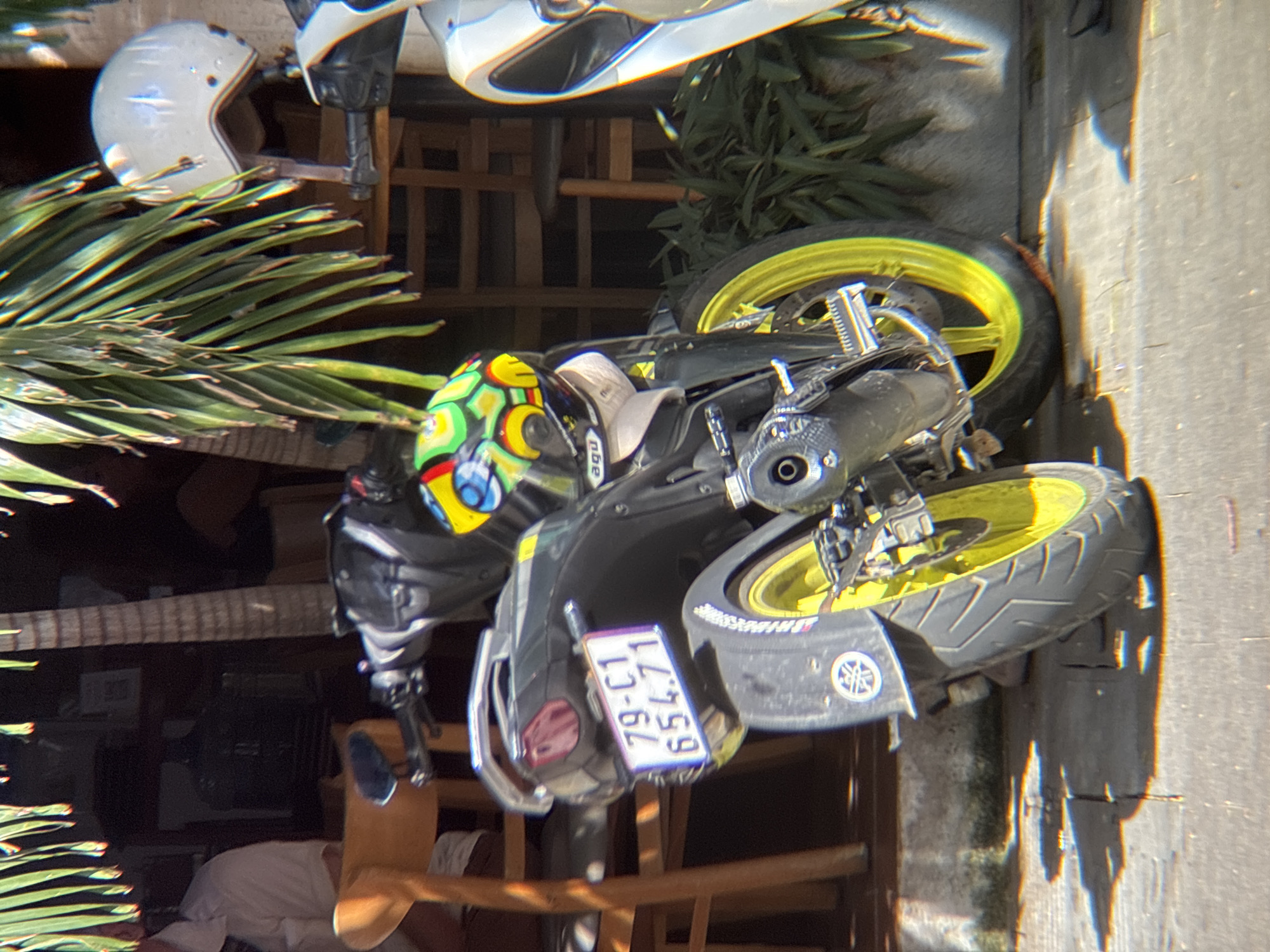 A motorcycle, with a license plate that reads "79-C1 654.71", is parked on a tiled surface in front of a building with a palm tree and other foliage. The motorcycle is black with yellow rims on its wheels and a colorful helmet is resting on its handlebars. A white helmet is visible on a shelf to the right of the motorcycle. The scene appears to be outdoors, possibly in a cafe or restaurant setting, in the city of Nha Trang, Vietnam, given the style of the motorcycle and the tropical vegetation. The lighting suggests it is daytime with strong sunlight creating shadows on the ground.