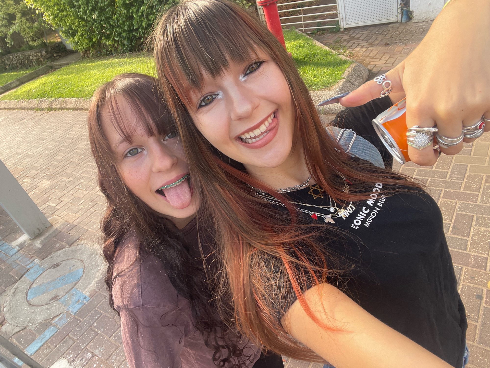 Two young women are posing for a selfie outdoors on a brick-paved walkway. The woman on the left has brown curly hair and is wearing a purple long-sleeved shirt. She is sticking her tongue out, which has green braces. The woman on the right has long brown hair with streaks of red and is wearing a black t-shirt with white text and multiple necklaces. She is smiling widely, her tongue also peeking out from her mouth, and she is holding an open can of what appears to be a beverage in her right hand. Her right hand is adorned with several rings. The background shows green foliage, a stone wall, a red post, and a white building with a fence. The image appears to have been taken in Raanana, Israel, possibly near a park or residential area, given the combination of greenery and paved walkways.