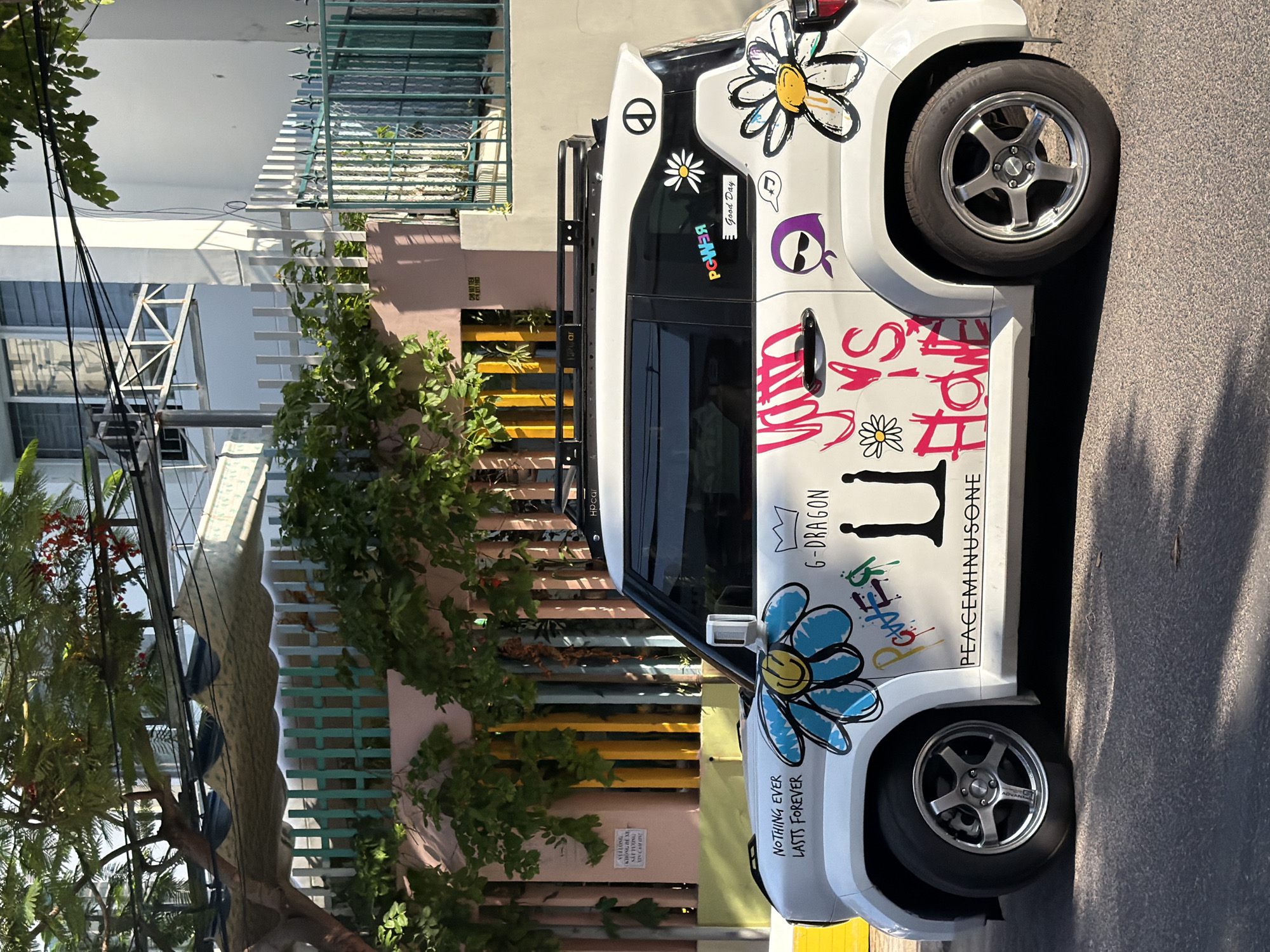 The image shows a white, four-door vehicle parked on an asphalt road in front of a pink and white building with a multi-colored fence and lush green trees. The car is heavily decorated with colorful drawings and text. On the driver's side, there's a large blue and yellow daisy with a smiley face, and text that reads "NOTHING EVER LASTS FOREVER". Above this, it says "G-DRAGON" with a crown symbol. Further back on the side of the car, there are more drawings including a daisy with a smiley face, a peace symbol, and the word "POWER" in colorful graffiti style lettering. There's also a sticker that says "Good Day". The rear of the car features a large, stylized daisy with yellow and orange petals and a black outline. The overall scene suggests a sunny day in a residential or commercial area in Nha Trang, Vietnam.
