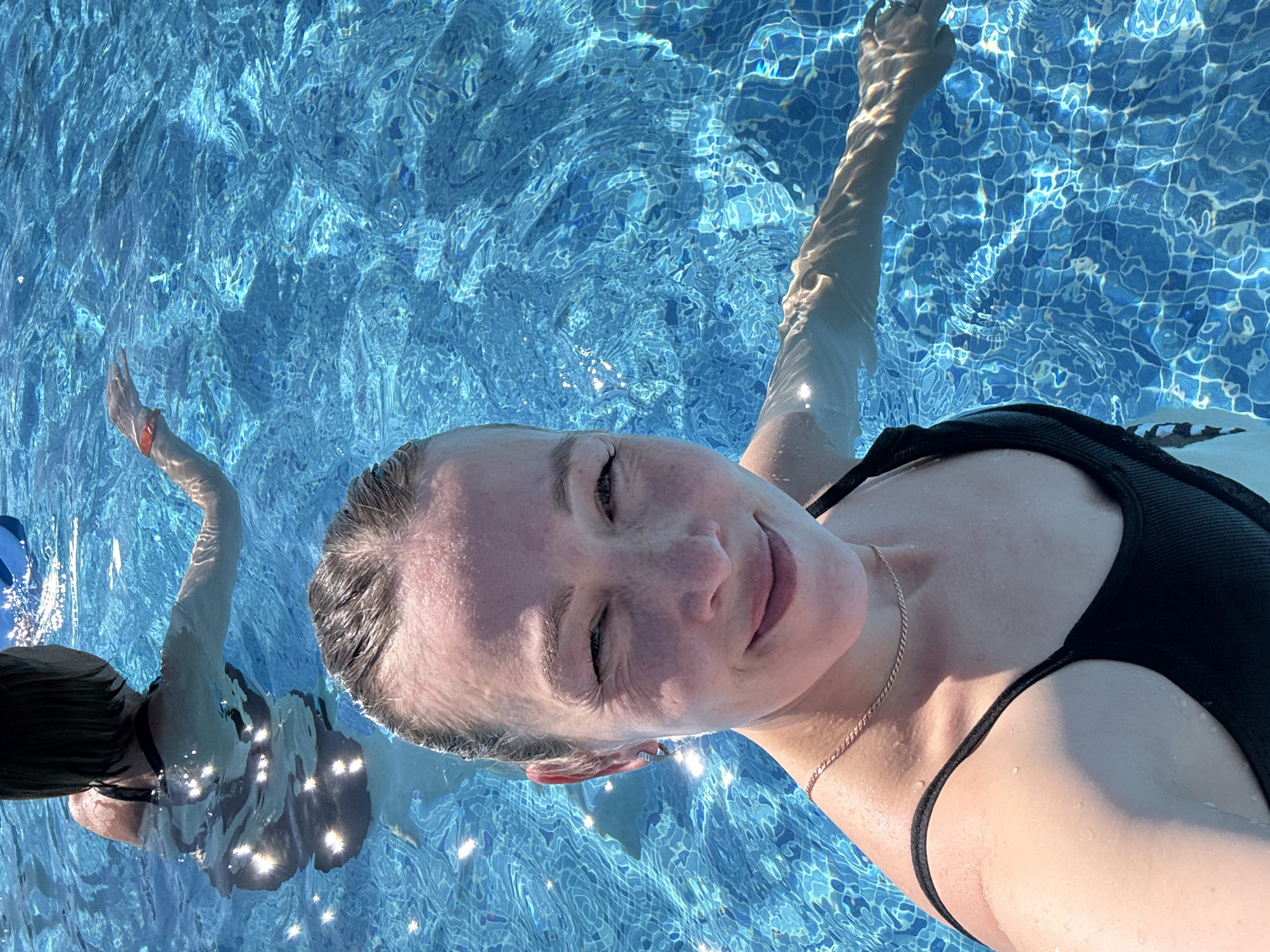 An overhead shot, slightly rotated, captures a young woman floating on her back in a vibrant blue swimming pool. She is looking upwards towards the camera, her eyes mostly closed, and a subtle smile on her face, suggesting relaxation or squinting in bright sunlight. Her dark hair is wet, and she wears a black swimsuit top and a delicate gold-colored necklace. Her arms are extended above her head, partially submerged in the water.
The pool water is clear and intensely blue, with strong sunlight creating shimmering, wavy patterns of light and shadow across the surface and onto the visible blue mosaic tiles of the pool floor. Water droplets are visible on her wet skin. A second person, also in the water, is partially visible in the background on the left, with an arm raised. The scene suggests a warm, sunny daytime, highlighting a tranquil moment in the water. The setting is a swimming pool, located in Votkinsk, Russia. No visible text is present.