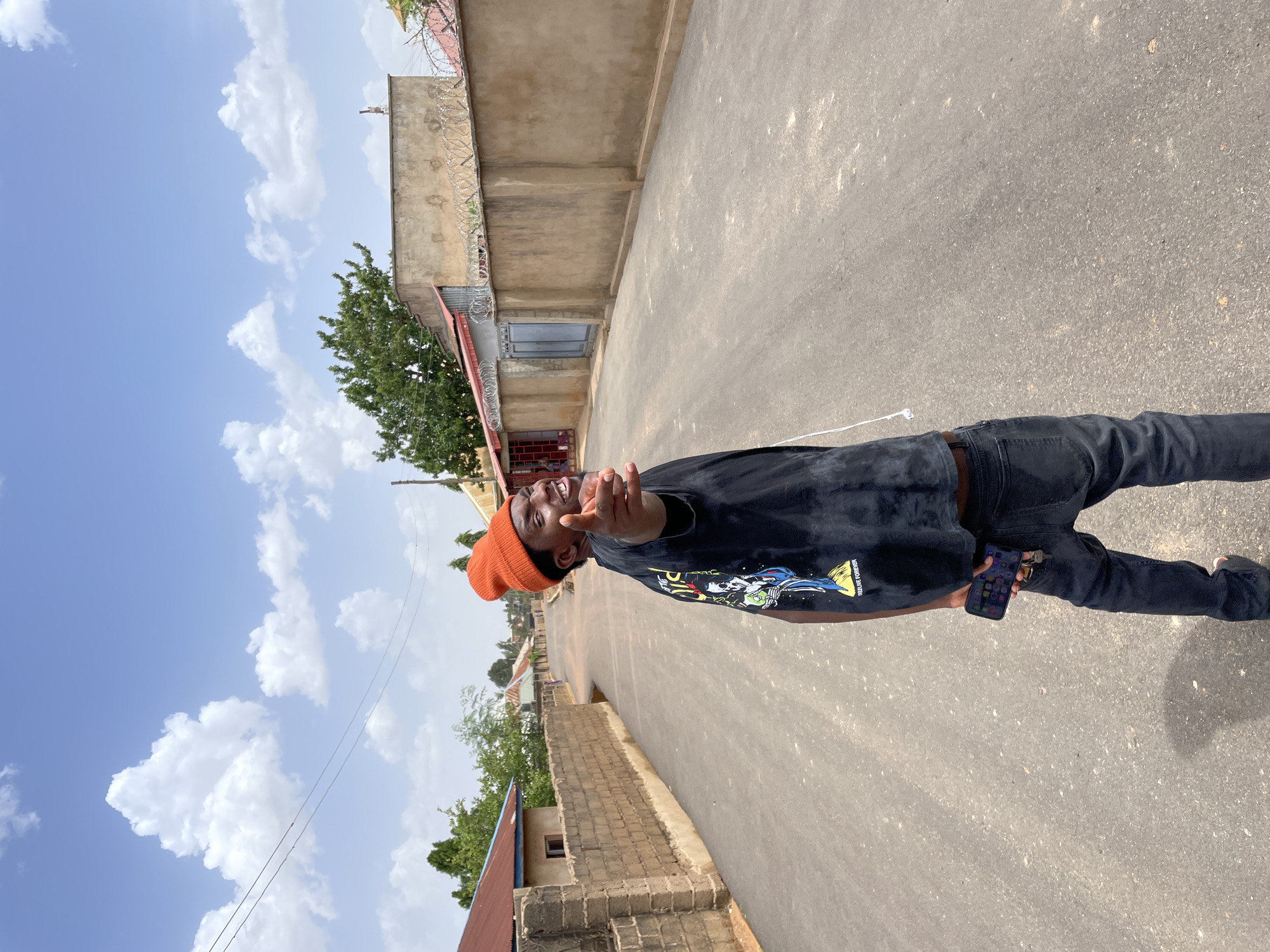 A full-length shot captures a young man standing on a paved road in Gura Top, Nigeria. He is wearing a dark-colored t-shirt with a graphic on the front, dark jeans, and an orange beanie. He holds a mobile phone in his left hand and gestures with his right hand towards the camera. The road stretches into the distance, flanked by buildings. On one side, a low brick wall runs along the road, while the other side features a taller building with a barbed wire fence on top. The sky above is bright blue, dotted with white clouds.