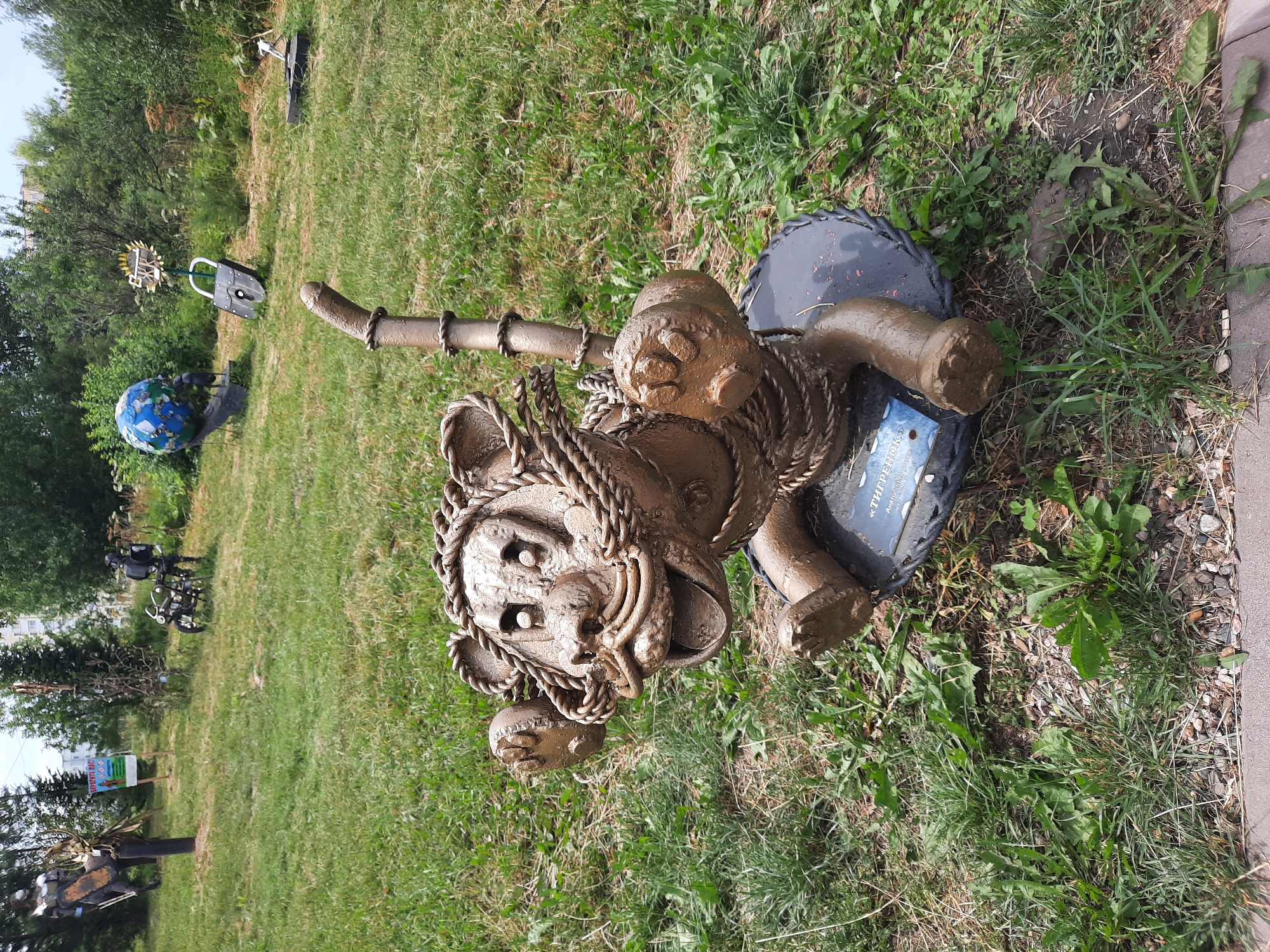 An outdoor metal sculpture of a cartoonish tiger sits on a dark circular base in a grassy area. The tiger sculpture is made of a rough, textured metal, painted a dull brown, and appears to be crafted from twisted ropes and pipes to form its body, limbs, head, and tail. Its eyes are large and hollow, and its mouth is open in a wide grin, revealing a tongue and teeth. It has large paws with visible claws and a long, segmented tail. A small blue plaque with white text is attached to the front of the circular base. The background consists of a moderately cut lawn with scattered weeds and small rocks. In the middle ground and background, other metal sculptures are visible, including a rider on a horse-like creature to the left, a motorcycle with a standing figure to the left-center, a globe on a pedestal, and a large padlock to the right. Trees and buildings are visible in the distance, suggesting an urban or park setting. The sky is overcast. The location is identified as Zelenogorsky, Russia.