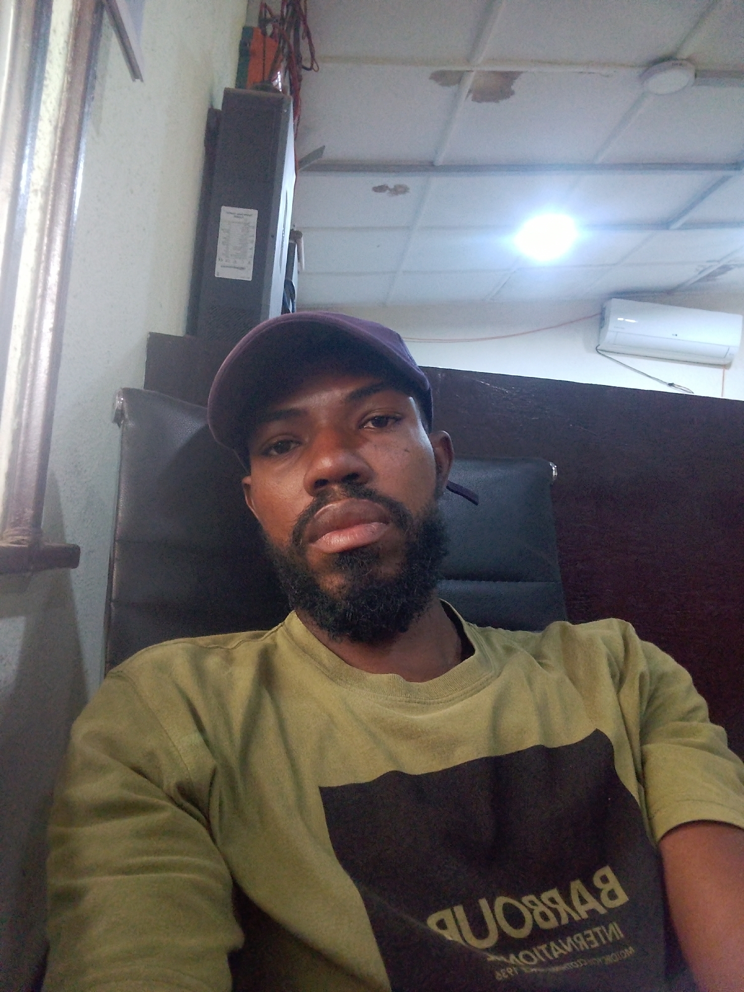 The media file is a selfie of a Black man wearing a light olive-colored long-sleeved t-shirt with a dark rectangular logo on the front. He is also wearing a dark purple baseball cap. The man has a beard and appears to be sitting in an office chair, with the back of the chair visible behind him. In the background, there is a white tiled ceiling with fluorescent lighting and an air conditioning unit mounted on the wall. A dark vertical structure with a label attached is visible on the left side of the image, possibly part of office equipment or a storage unit. The overall setting suggests an indoor environment, likely an office or workspace, in Palmgrove, Nigeria. The lighting appears to be artificial, indicating it could be daytime or evening. The man's expression is neutral, and there are no overt indications of any specific activity or event. The text on the t-shirt logo is partially visible and appears to be in a lighter color, possibly yellow or white. It seems to read "BARBOUR INTERNATIONAL 1936" in reverse, suggesting it might be a mirrored reflection or intentionally reversed text.