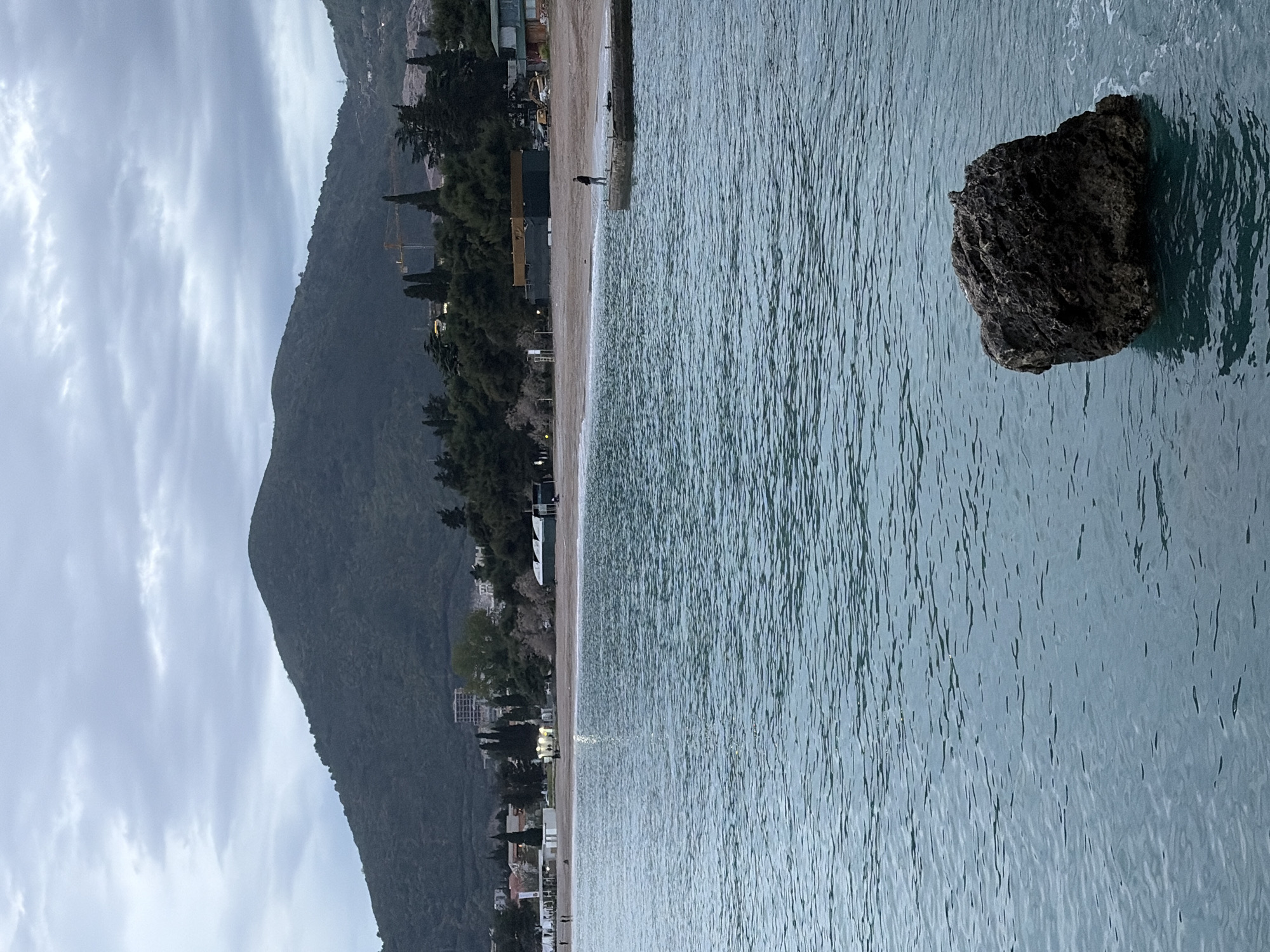 The image captures a scene in Budva, Montenegro, featuring a view of the coastline. A large mountain dominates the left side of the frame, gradually descending towards the coastline. A sandy beach runs along the edge, lined with trees and buildings. A person is standing on a concrete pier that extends into the water. The water's surface reflects the sky and exhibits a ripple pattern, indicating a slight breeze. A large, dark rock sits partially submerged in the water on the right side. The sky is overcast with a layer of clouds.