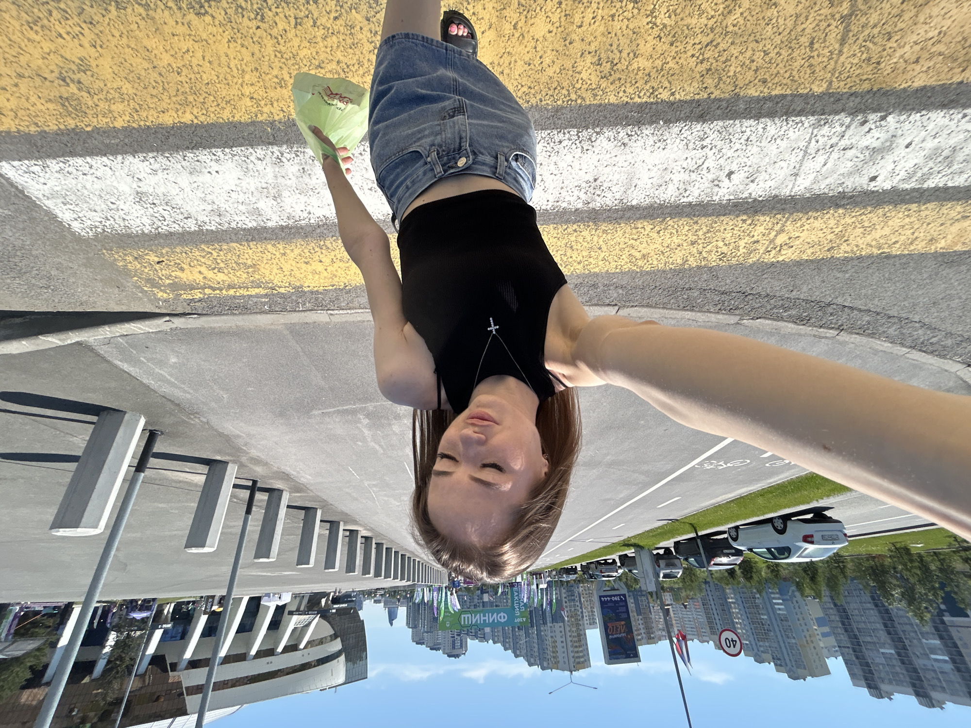 A young woman is captured in a selfie on a bright, sunny day, positioned slightly left of center and looking down at the camera held by her outstretched right arm. She wears a black ribbed crop top, a light blue denim mini skirt, and dark sandals. A silver cross necklace adorns her neck, and she holds a light green plastic bag in her left hand. She is walking on a zebra crossing with alternating yellow and white stripes. To her immediate right, a wide asphalt pedestrian path is bordered by a row of gray hexagonal concrete bollards and several tall, slender light poles, leading towards a large, modern, multi-story stadium with a curved white and glass facade. To her left, beyond a grassy median and a designated bike lane marked with a bicycle symbol, an asphalt road features two lanes of traffic, including a white Skoda Rapid, a dark sedan, and a white SUV. A circular "40" speed limit sign is visible above the road. In the background, a row of distinctive modern high-rise residential buildings with angled facades extends, interspersed with event banners displaying Russian text such as "ФИНИШ" (FINISH) and "АЗОТ РЕАКЦИИ" (NITROGEN REACTION). The environment is an urban setting under a clear blue sky, identified as being in Moscow, Russia, near the VTB Arena – Dynamo Central Stadium.