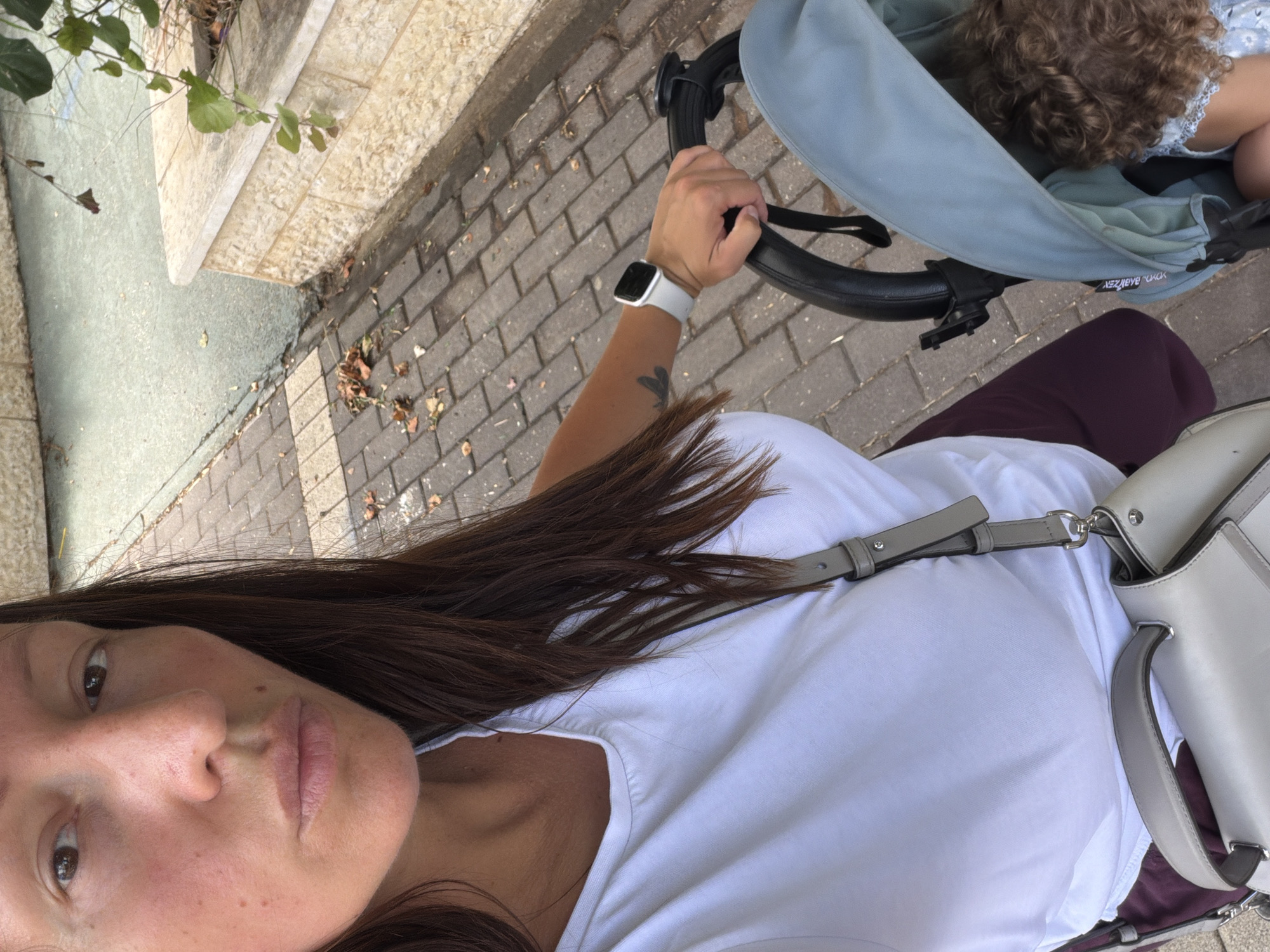 A woman is seen from the chest up, pushing a baby stroller on a brick sidewalk. She has long, dark brown hair and is wearing a white t-shirt. A light-colored purse is slung over her shoulder. Her left hand is on the stroller handle, and she is wearing a white smartwatch. The stroller is light blue and contains a baby with curly brown hair. The background shows a light-colored stone wall and some greenery. The scene appears to be in an urban environment, possibly a street or park.