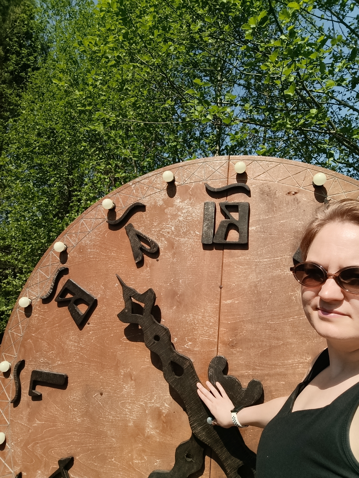 A person stands to the right of a large, outdoor clock in Prikamskii, Russia. The clock face is brown with dark, stylized numerals and ornate hands. Small, white bulbs are evenly spaced around the clock's perimeter. The person, wearing sunglasses and a black tank top, has their hand resting on the clock's minute hand. Green trees fill the background, with patches of blue sky visible through the branches.
