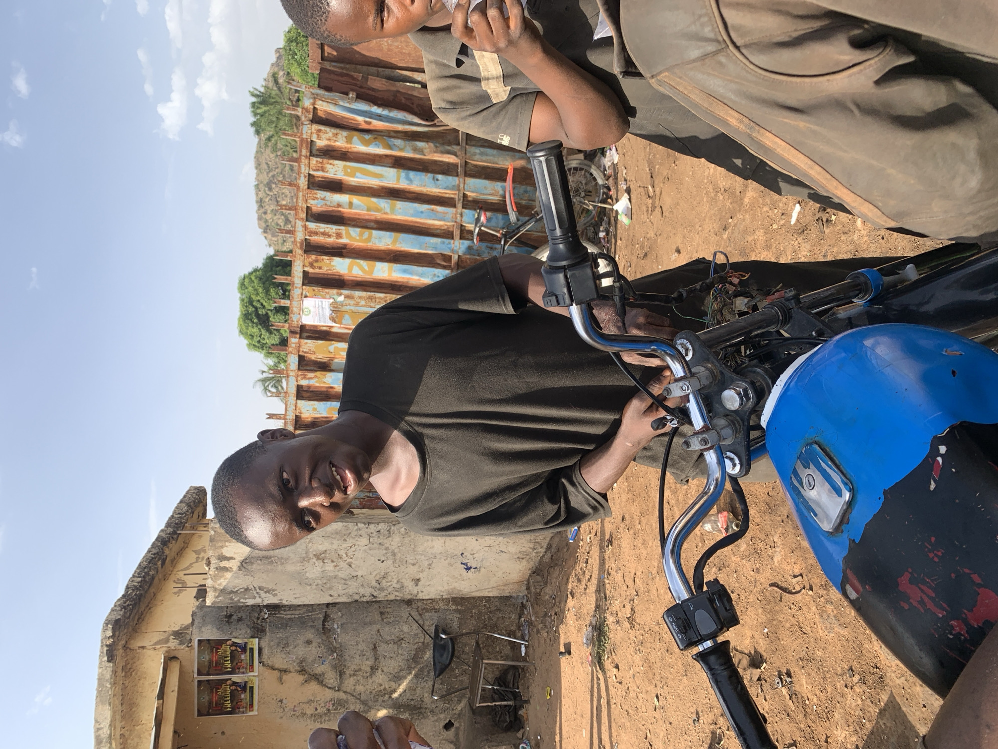 The image captures a scene in Gura Top, Nigeria, where a man is focused on repairing the electrical components of a motorcycle. He is dressed in a dark green T-shirt, working on the bike's wiring near the handlebars. Another person is partially visible next to him. The motorcycle has a blue and black fuel tank and chrome handlebars. The setting includes a weathered building with posters on the wall, a rusty corrugated metal fence, and a dirt ground. A glimpse of a hill or small mountain is visible behind the fence.