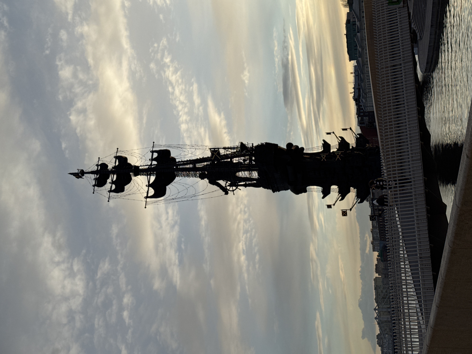 The image shows a silhouette of the Peter the Great Statue in Moscow, Russia. The monument depicts a large ship with sails against a cloudy sky. The ship is seen from a low angle, possibly from a bridge, as a section of the bridge's railing is visible on the right side of the frame. Buildings can be seen in the background, indicating an urban environment. The scene suggests the statue is located near the water, likely the Moskva River.