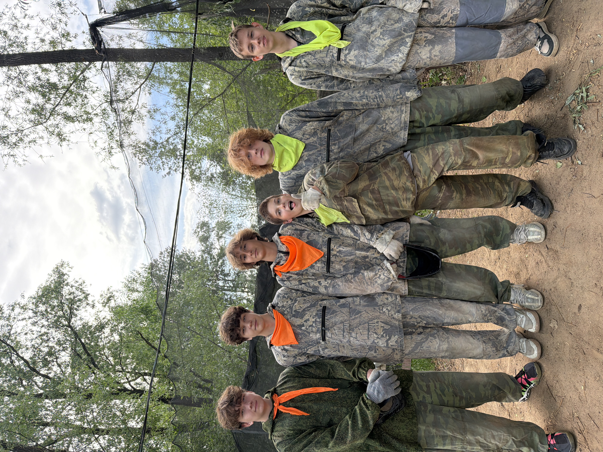 A group of six young individuals stand in a line outdoors, likely at a paintball or similar recreational facility, potentially located in the city of Chaikovsky, Russia. All individuals are wearing camouflage clothing, with each wearing either a bright yellow or orange bandana or neckerchief. The attire suggests they are participants in an activity such as paintball or airsoft. The background includes trees and a net structure overhead. The ground appears to be dirt and vegetation. The sky is visible with clouds.