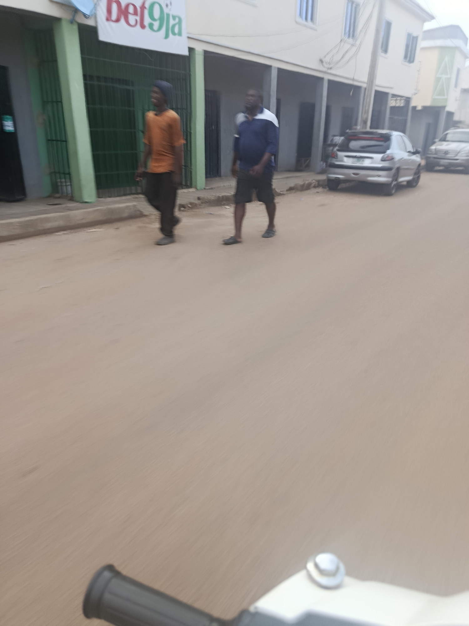The image captures a street scene in Rafin Par, Nigeria. Two men are walking on a paved road. The man on the left wears an orange t-shirt and dark trousers. The man on the right wears a blue and white polo shirt and dark shorts. Buildings line the left side of the street, one of which displays a "Bet9ja" advertisement. Cars are visible parked on the right side of the street. The perspective suggests the photo was taken from a moving vehicle, possibly a motorcycle, as indicated by the blurry handle in the foreground. The overall environment appears to be a typical urban street with moderate pedestrian and vehicular activity.