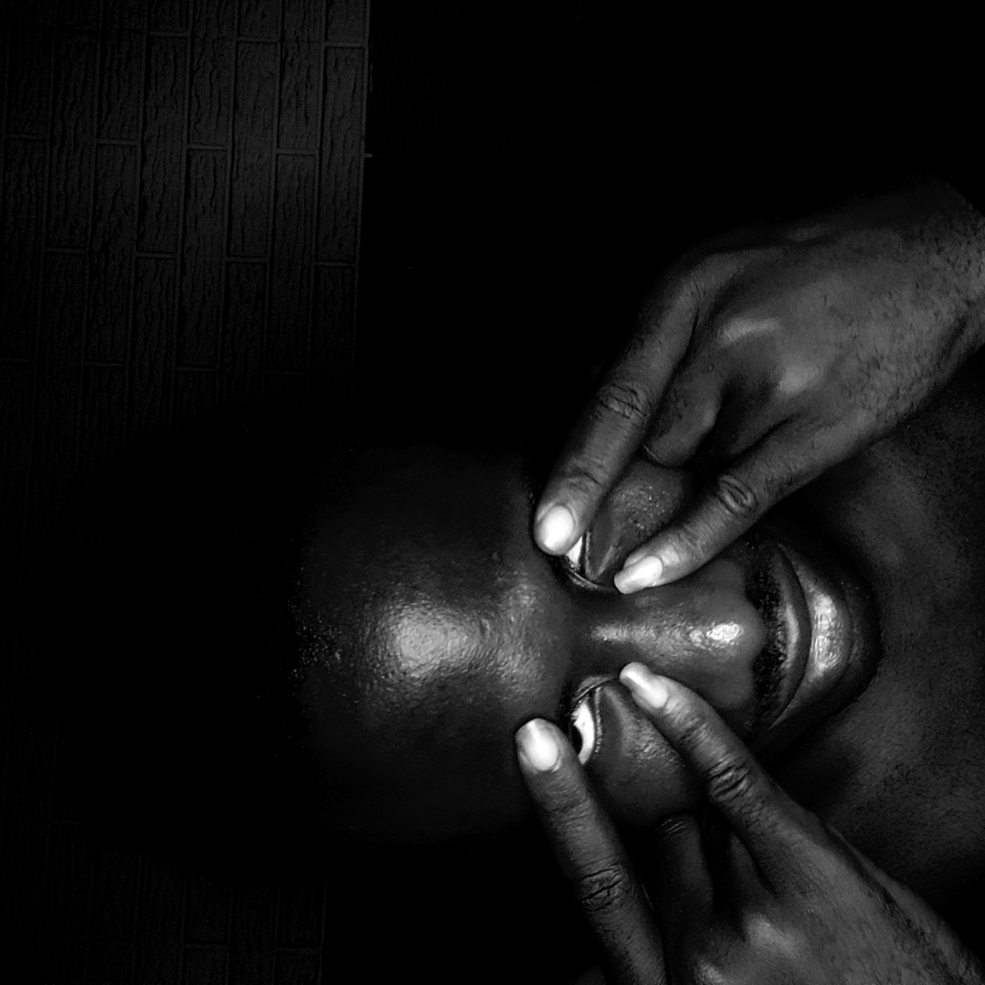 The image is a black and white close-up photograph. It shows a person lying down, possibly on their back, with two hands covering their eyes. The person's face is visible between the fingers of the hands. The skin is dark and appears slightly glossy. A textured wall is visible in the upper left corner of the frame. The rest of the background is dark and out of focus. There is no specific information in the photograph to suggest it was taken in Narayi, Nigeria.