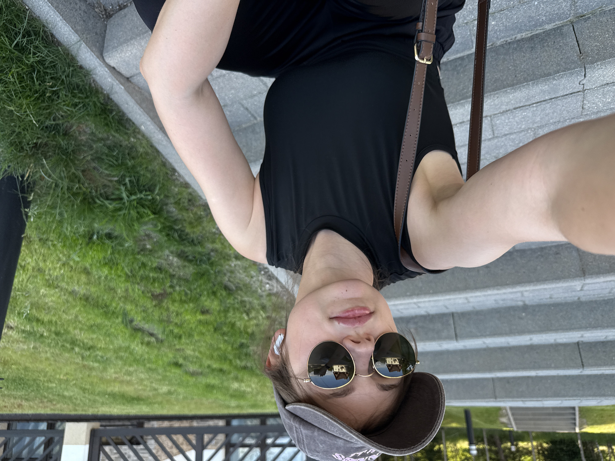 A young woman wearing a black tank top, black pants, a gray baseball cap, round sunglasses, and white wireless earbuds sits on stone steps next to a grassy area. A brown purse strap is visible over her left shoulder. The sunglasses reflect two houses. The background shows a pathway with more steps, trees, and a fence. The scene appears to be outdoors in a park or public area. The location is not specified in the image itself, and there is no information to suggest it is in Szklarska Poręba, Poland.