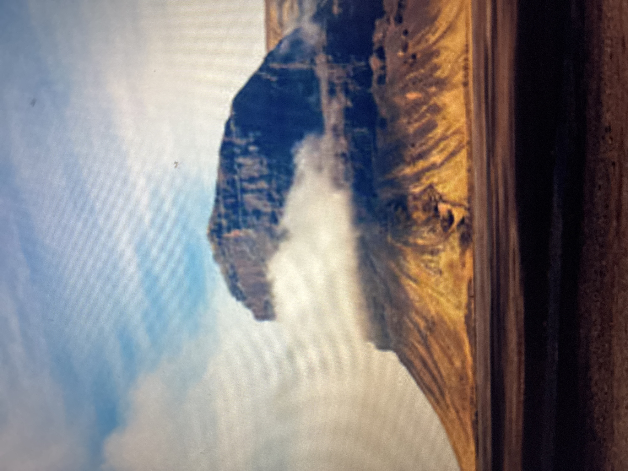 The image displays a rugged, misty mountainous landscape, likely a natural landmark near Asenovgrad, Bulgaria, given the provided context. The photograph itself appears to be of a screen, showing a visible pixel grid across the entire frame.
In the left portion of the frame, a large, dark, imposing mountain or rock formation dominates, rising steeply. Its dark, textured surface, possibly composed of cliffs, is partially shrouded in significant white mist or low-hanging clouds, particularly around its base and mid-section, which creates a dramatic and atmospheric effect.
To the right, a contrasting, lighter-toned mountainous or hilly terrain extends across the midground and foreground. This area is characterized by earthy colors, primarily shades of brown and ochre, with visible undulating textures and patterns that suggest eroded slopes or sparse vegetation. The prevalence of mist also affects the clarity of this terrain.
The upper part of the image reveals a bright sky with patches of softer white clouds, indicating a daytime setting. However, the extensive mist and cloud cover around the mountains suggest a cloudy or overcast day, possibly with cool or humid weather conditions.
There are no visible people, identifiable man-made objects beyond the natural landscape, or discernible text within the scene. The overall impression is one of majestic natural beauty and solitude.