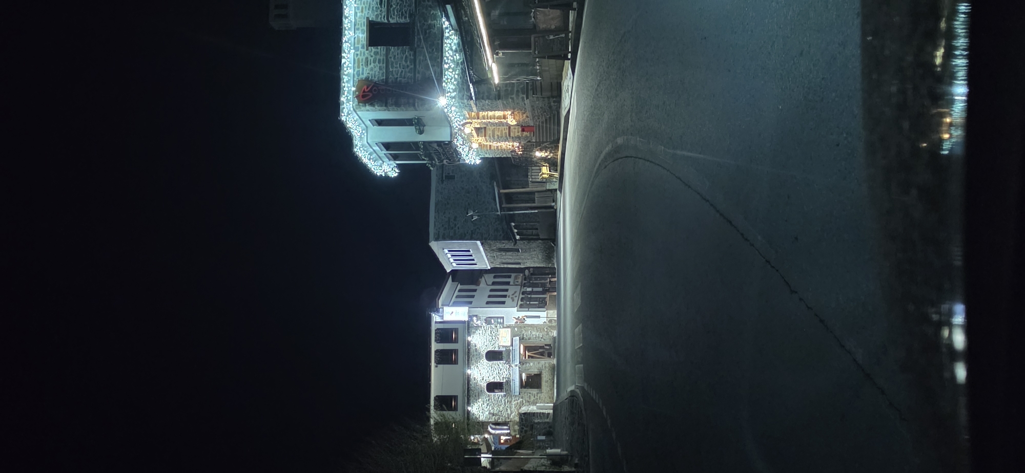 The image shows a night scene in Shiroka Laka, Bulgaria. The street is dark with a cobblestone road curving to the right. Stone buildings line the left side of the street, some decorated with Christmas lights. No people are visible. The focus is on the architecture and the festive lighting.