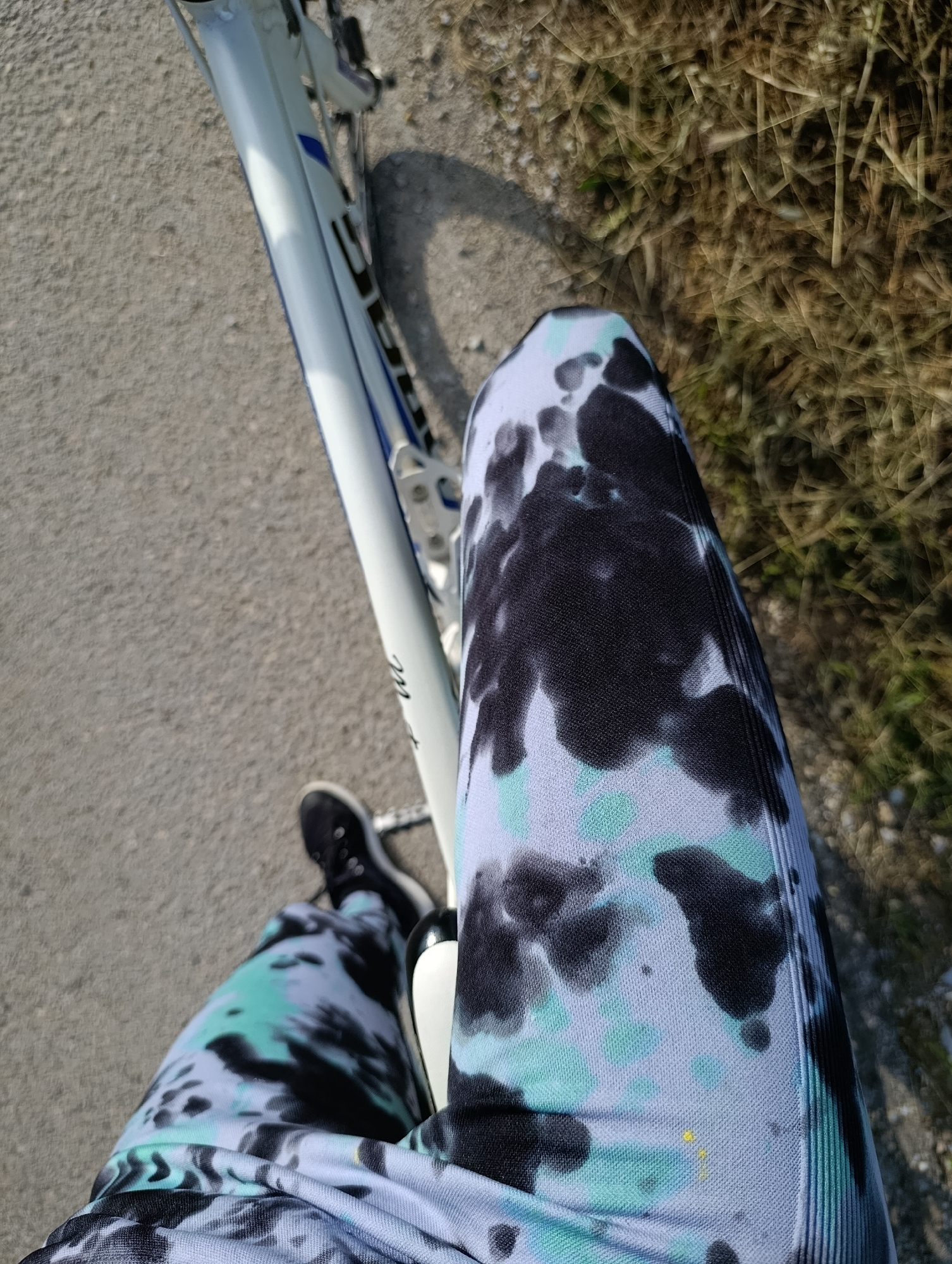 A first-person perspective photo taken outside, likely near Generalovo, Bulgaria. The subject is sitting on a bicycle on a paved road next to dry grass. The bicycle is white with blue accents. The person is wearing black sneakers and tie-dye leggings with black, white, and light blue.