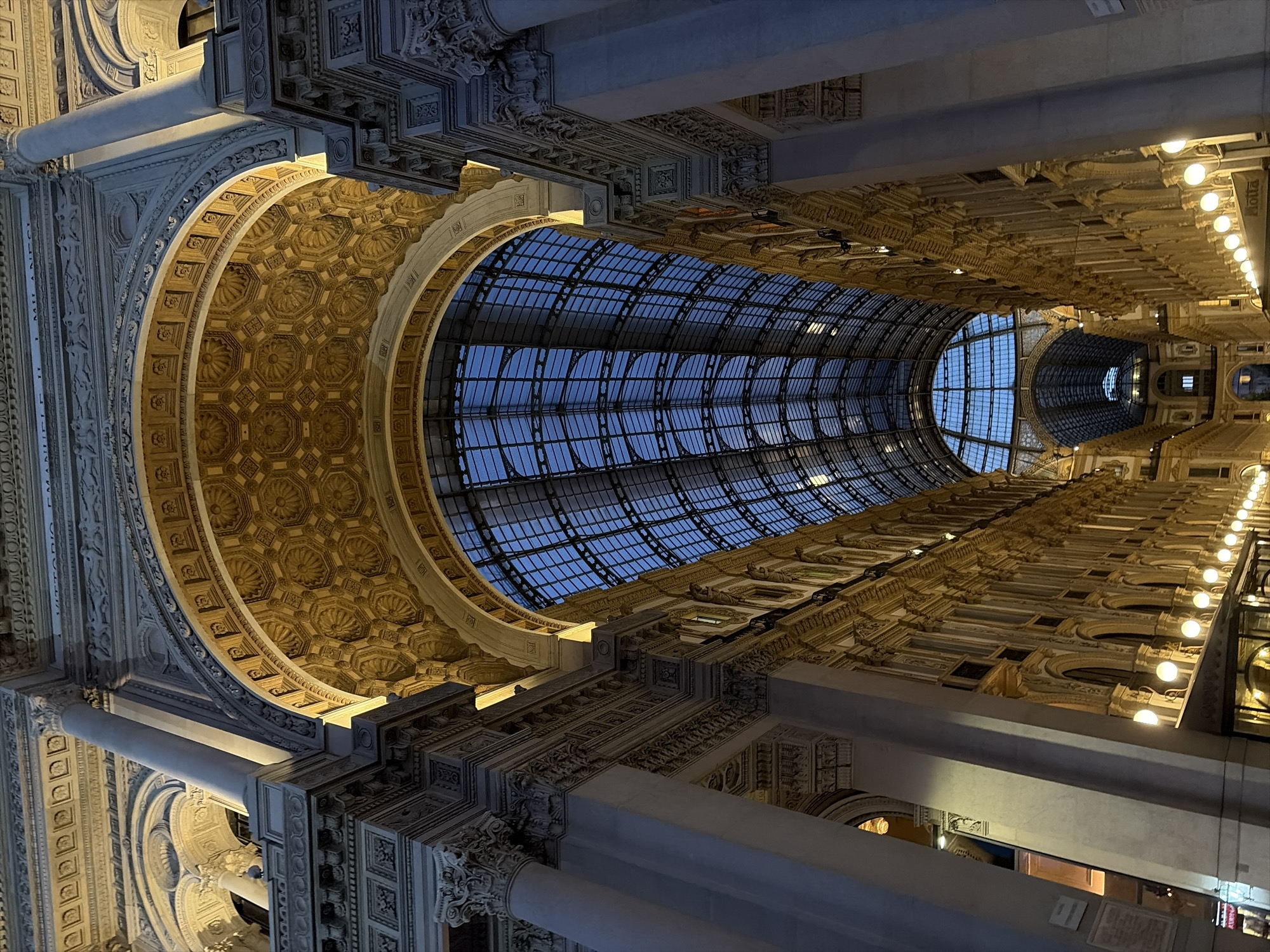 The image shows the Galleria Vittorio Emanuele II in Milan, Italy, at dusk. The grand arched structure is illuminated by artificial lights, highlighting the ornate architectural details. A glass and iron roof spans the length of the Galleria, revealing a deep blue sky above. Rows of shops with ornate facades line both sides of the arcade. The perspective is looking up and down the length of the Galleria, emphasizing its vastness and architectural splendor. No people are clearly visible in the image.