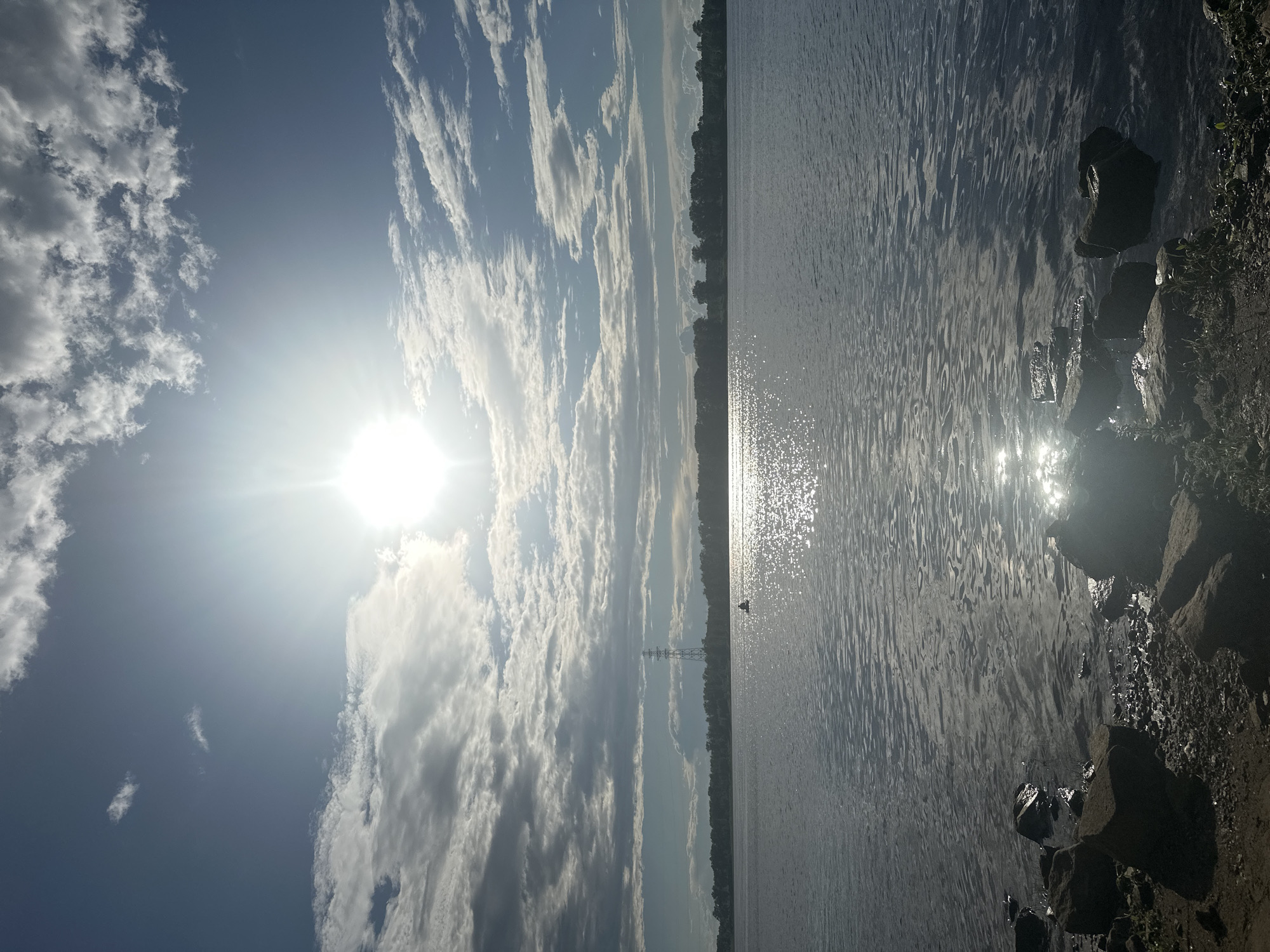 This image depicts a serene waterscape on a sunny day. The sun, positioned high in the sky to the left, casts a bright glare and creates shimmering reflections on the water's surface. Scattered white clouds dot the clear blue sky. The water body appears to be a large lake or river, with a distant treeline marking the opposite shore. A small boat is visible in the middle distance on the water. The foreground shows a rocky shoreline with some vegetation. The time of day is clearly daytime, with the bright sun indicating it could be mid-morning or afternoon. The weather is fair and sunny. The scene evokes a sense of peace and natural beauty. There is no visible text in the image. The location context suggests this scene is in Chaikovsky, Russia.