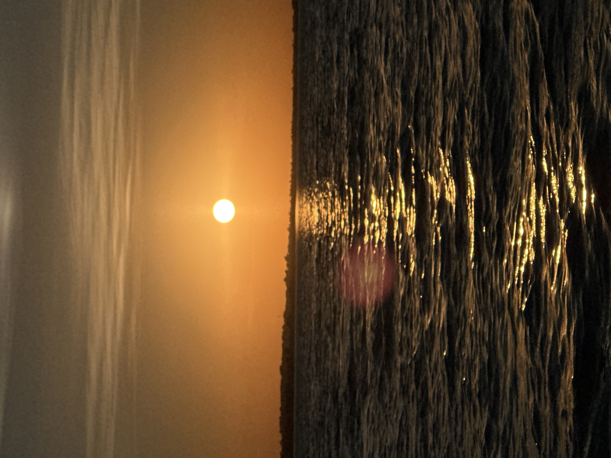 The image depicts a serene sunset over a body of water, likely a lake, with a distant treeline on the horizon. The sun, a bright orb, is positioned in the upper center of the frame, casting a warm orange glow across the sky and reflecting off the rippling water. The sky is streaked with thin clouds, adding texture to the scene. The water is dark and choppy, with streaks of golden light shimmering on its surface. No people or specific landmarks are visible in the image. The scene is evocative of a natural landscape at dusk.
