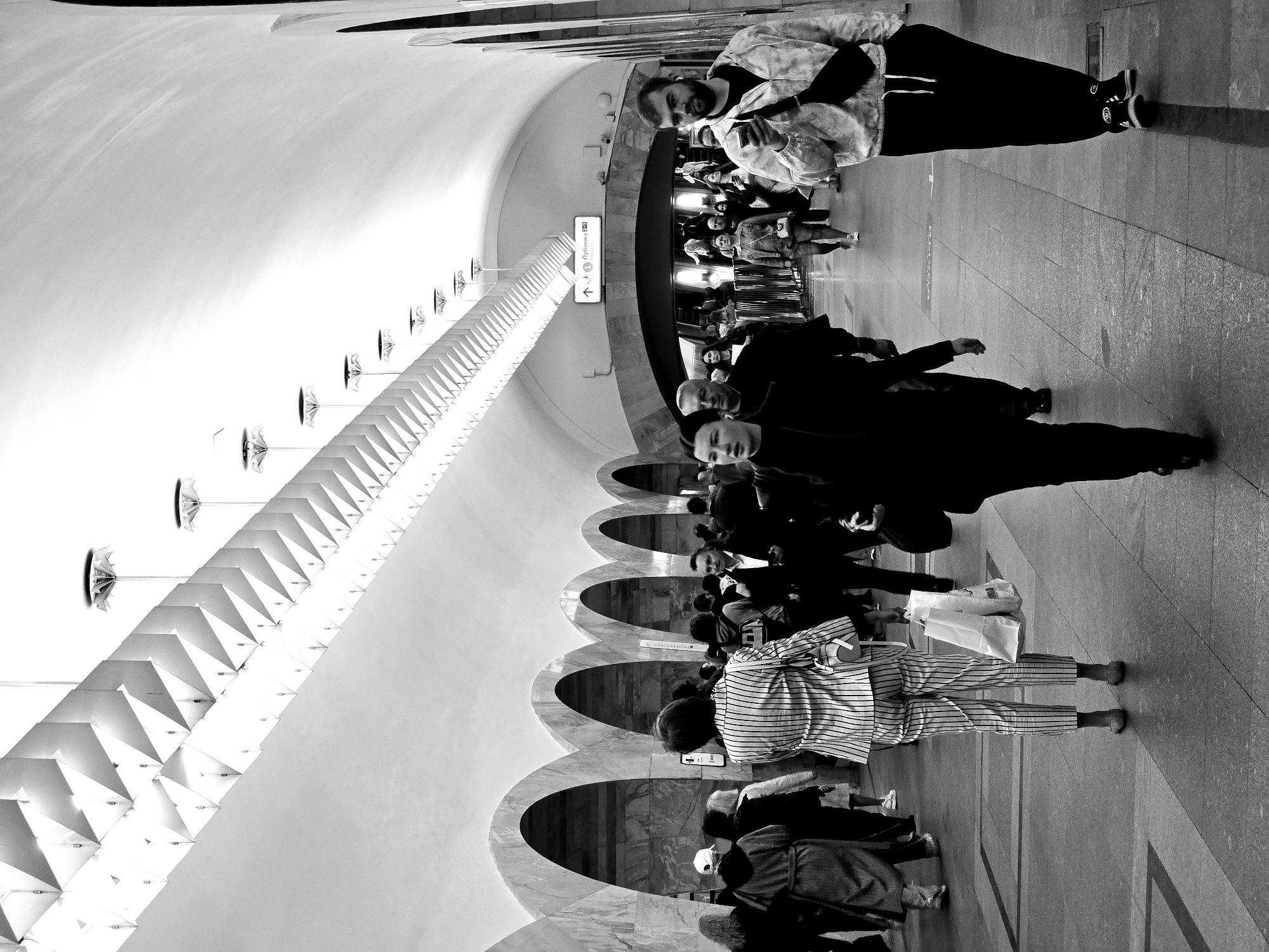 The image shows a black and white view of an underground train station, most likely the Moscow Metro in Russia. The station features an arched ceiling with a series of repeating arches along the side. A row of triangular fixtures and umbrella-like lamps runs along the top of the wall. A crowd of people is walking along the platform, in different directions. A man in a tie-dye jacket is looking at his phone. A sign in Russian with a directional arrow pointing left is visible on one of the arches.