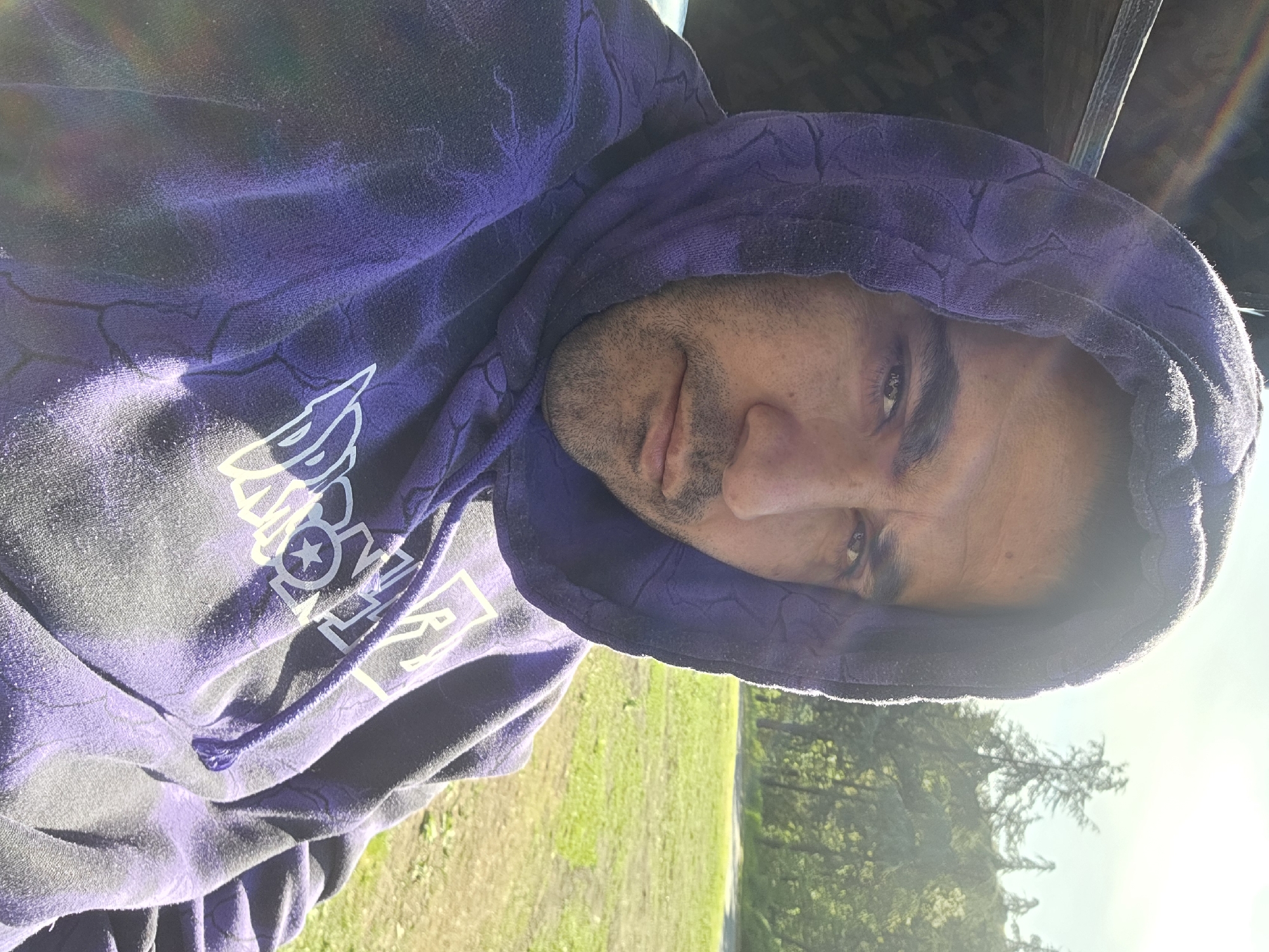 A man is wearing a purple hoodie with white lettering and a hood. The hoodie has a crackle pattern. The background shows green grass and trees illuminated by sunlight.
The location is not discernible from the image. There is no evidence to indicate that this image was taken in Chargan, Bulgaria.