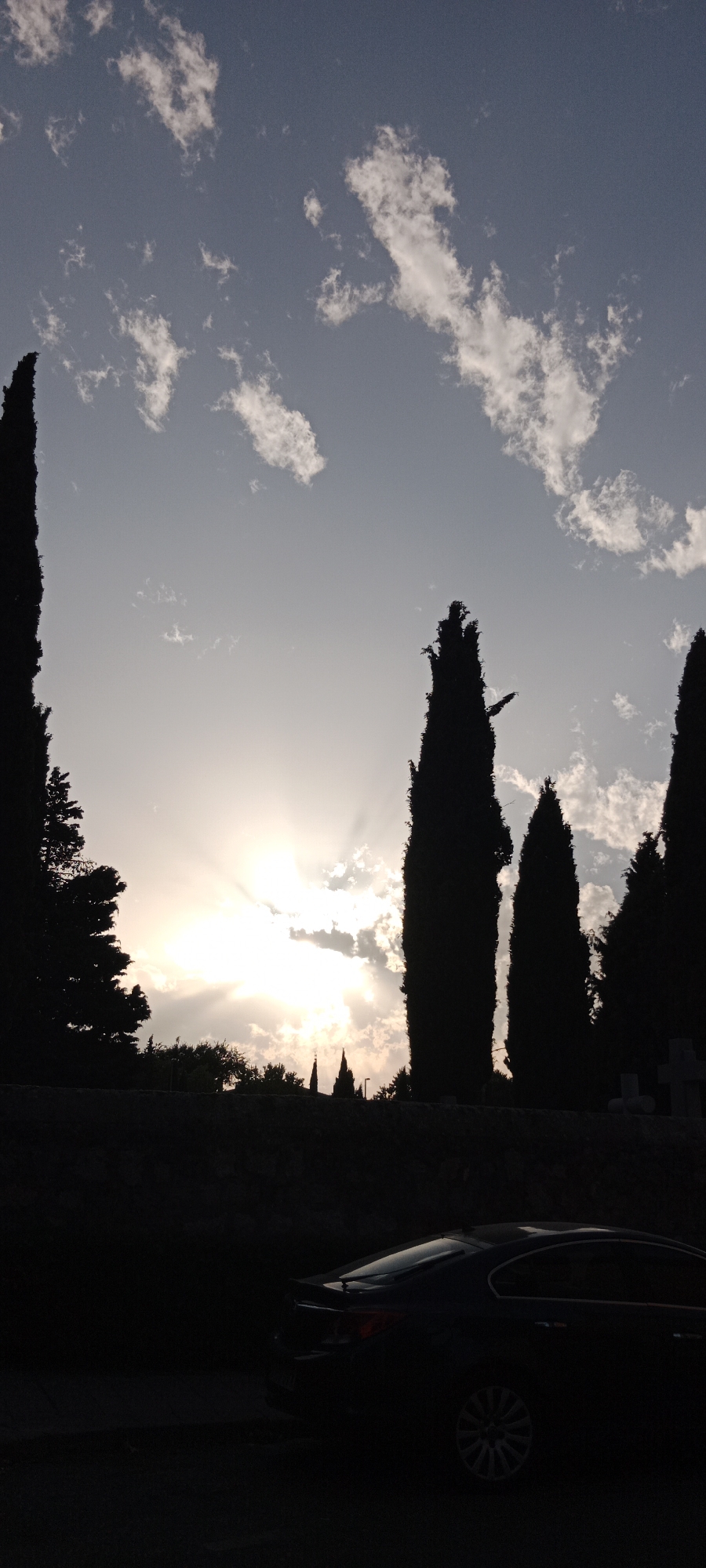 The image shows a cemetery in Galapagar, Spain, at dusk. Tall cypress trees line the background, silhouetted against a sky filled with scattered clouds and the setting sun. The sun's rays pierce through the clouds, creating a dramatic effect. In the foreground, a dark-colored sedan is parked, its features mostly obscured by shadow. The overall scene is one of quiet solemnity, with the natural beauty of the twilight sky contrasting with the somber atmosphere of the cemetery.