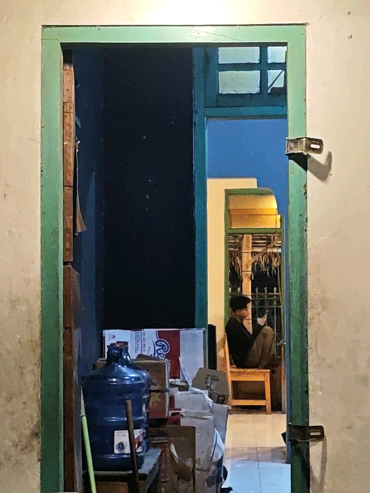 The image, captured in Ngasem, Indonesia, presents a view through an open, weathered green door frame into a dimly lit interior and an adjacent outdoor space. In the foreground, to the left, a large blue water jug is positioned next to some cardboard boxes, one with red and white branding. The wall to the right of the door frame is a pale, textured color, with a metal latch affixed to it.
Beyond the door frame, the immediate interior is dark. Further back, through a second, darker green doorway with a small, four-paned window at the top, a young male is seated on a simple wooden chair in what appears to be an outdoor or semi-enclosed area. He is wearing a dark long-sleeved shirt and camouflage-patterned trousers, and is focused on a mobile phone he holds in his hands. The background behind him includes a barred window and some hanging items. The overall lighting suggests it is either dusk or nighttime, with artificial light casting a warm glow from the area where the person is seated. The scene conveys a sense of domestic or utilitarian space, with evidence of everyday objects and the quiet activity of an individual.