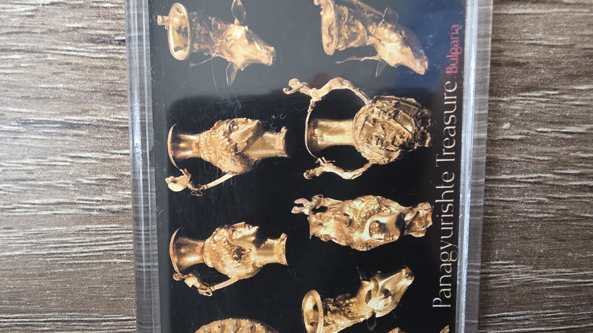 The image shows a close-up view of the cover of a cassette tape. The cover features a black background with a pattern of golden Thracian rhytons, vessels used for drinking, from the Panagyurishte Treasure. The words "Panagyurishte Treasure Bulgaria" are printed vertically on the right side of the cassette cover. The cassette tape is encased in a clear plastic case. The tape, representing cultural treasures, is not directly linked to Blagoevgrad, Bulgaria, but rather Panagyurishte.