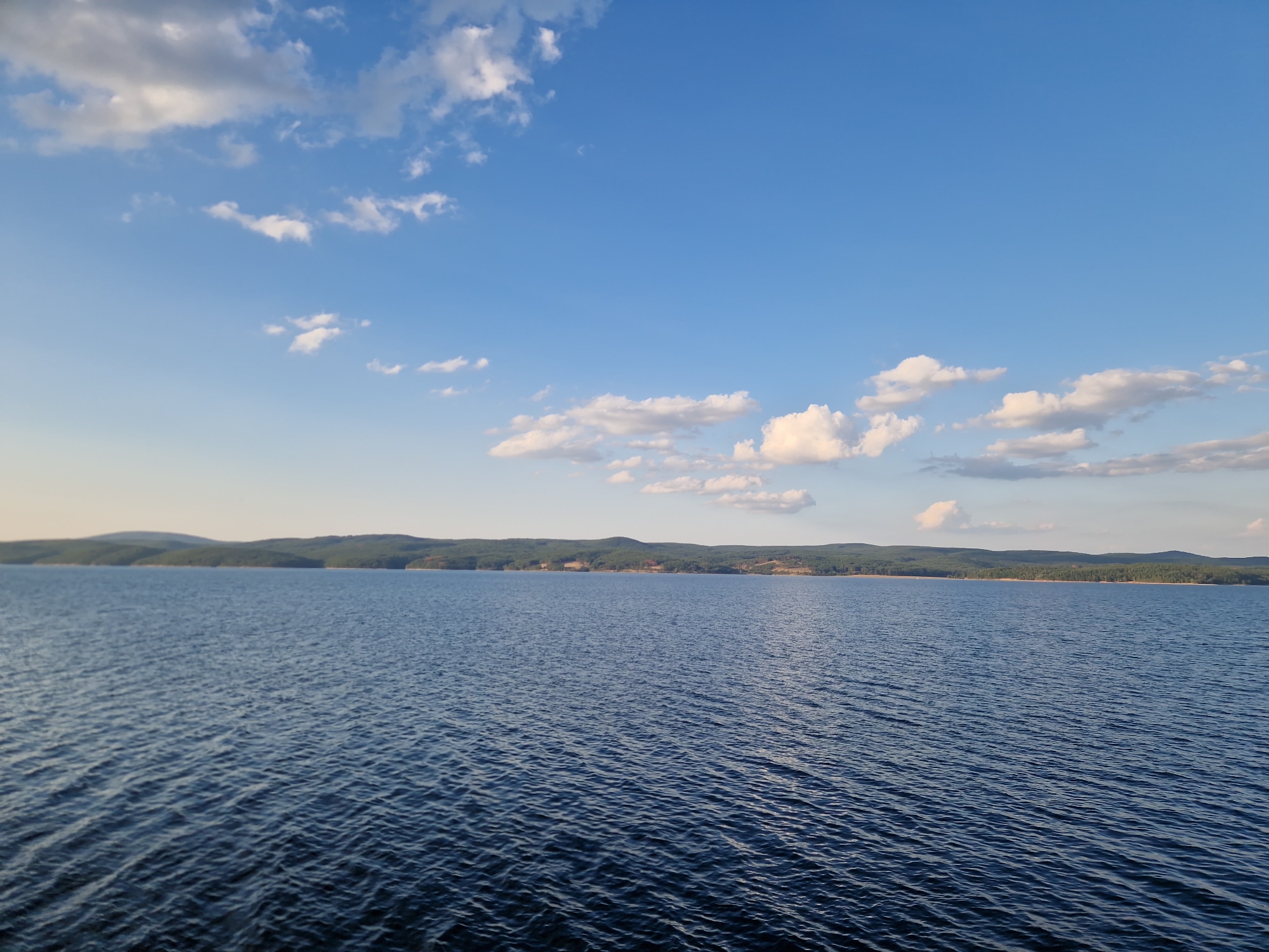 A wide shot captures a calm, dark blue body of water extending towards a distant, tree-covered shoreline under a bright blue sky with scattered white clouds. The water's surface is rippled, reflecting the sky. The shoreline is a continuous band of green, with gentle, rolling hills forming the backdrop. The scene appears to be a natural landscape, possibly a reservoir or lake, situated in or near Dolni Pasarel, Bulgaria, given the context. No people or man-made structures are immediately visible on the water or the shoreline in this frame. The overall impression is one of serene natural beauty.