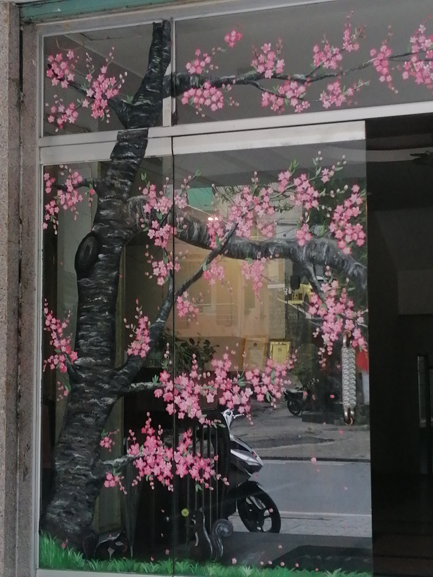 The image depicts a mural of a cherry blossom tree painted on a glass storefront in Da Lat, Vietnam. The mural features a large, dark tree trunk with branches adorned with abundant pink blossoms. The painting extends across multiple glass panes, creating a continuous scene. Reflections of the street, including a parked motorcycle and buildings, are visible on the glass. The bottom of the mural depicts green grass and what appear to be black cats. The overall impression is of a vibrant, artistic decoration enhancing the storefront.