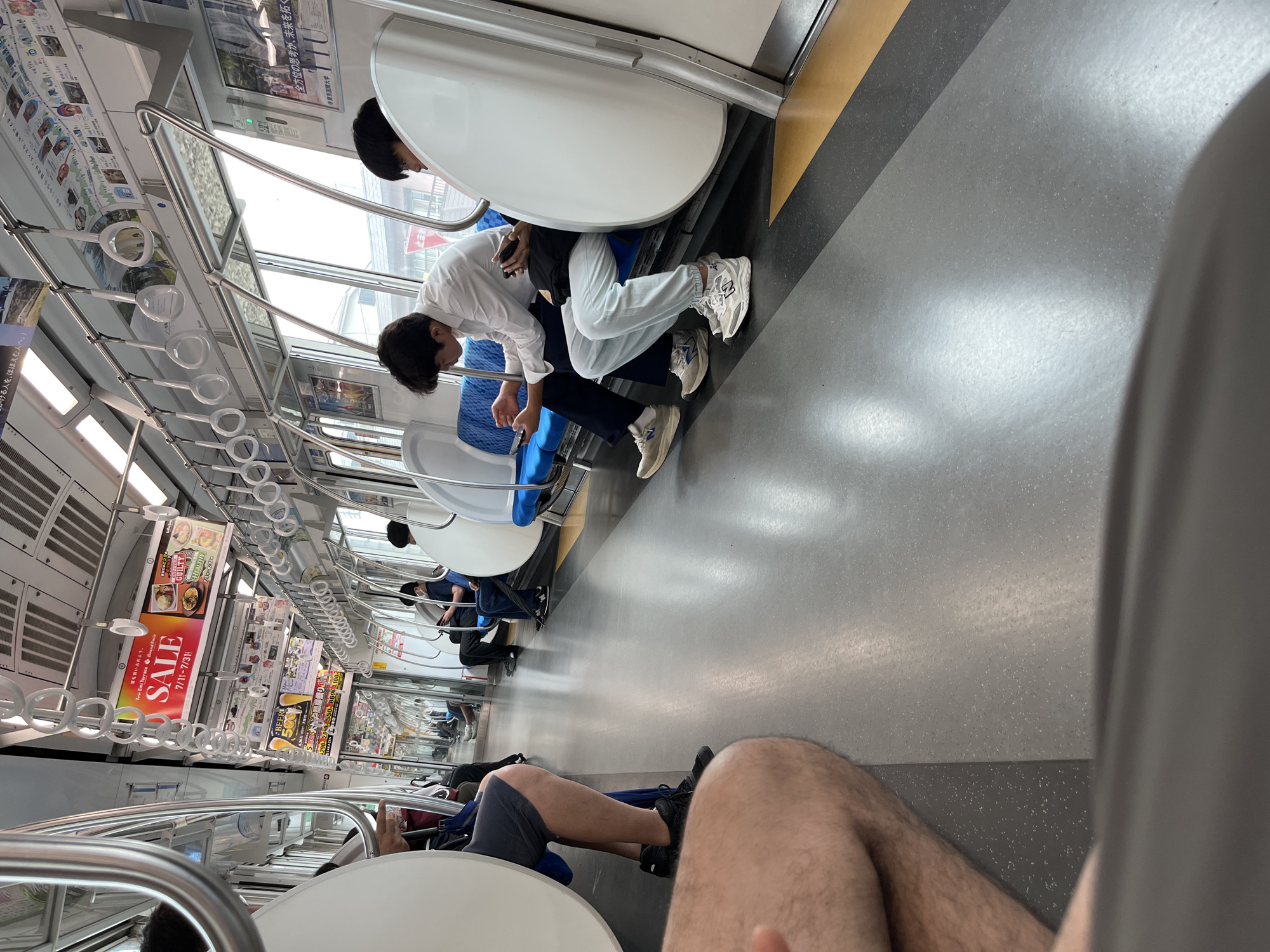 The image depicts the interior of a commuter train with passengers seated. The train appears to be in motion, with reflections visible on the floor and windows. The scene is set on a train in the city of Sayama, Japan.
In the foreground, a person's leg and thigh are visible, covered in hair, suggesting they are seated on the left side of the train. The floor is a muted gray with some texture and is slightly reflective, showing streaks of light and shadow.
Further down the aisle, two young men are seated on blue upholstered seats. The man on the left, wearing a white long-sleeved shirt and dark pants, is looking at his phone, which he holds in his right hand. He is wearing light-colored sneakers. The man to his right, wearing a dark top and light-colored pants, is also looking at his phone, which he holds in his left hand. He is wearing light-colored sneakers with blue accents.
Other passengers are visible further back in the train, some standing and holding onto handrails. The train's interior features numerous handrails for standing passengers, and advertisements are displayed above the windows. The windows show a blurred view of the outside, indicating movement. The overall environment is clean and utilitarian, typical of public transportation.
