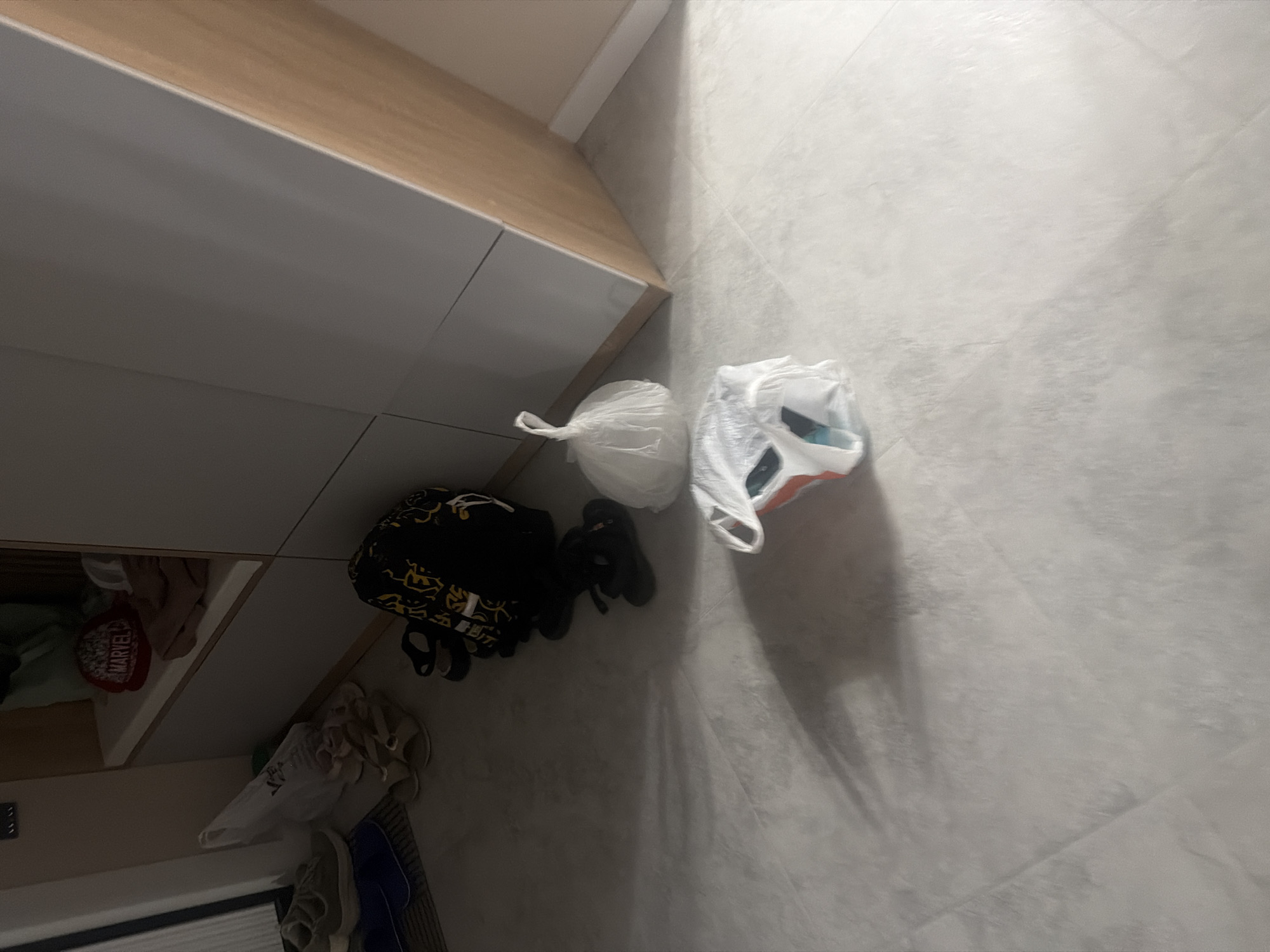 The image depicts an indoor scene, likely a hallway or entryway, with light-colored tiled flooring. Several items are scattered on the floor, suggesting a temporary or disorganized state. To the left, a pair of green athletic shoes rests on a dark mat. Adjacent to these are two pairs of sandals, one beige and one dark. In the center of the frame, a black backpack with a yellow graphic pattern is visible, along with a pair of black flip-flops. Further to the right, two white plastic bags are present; one is a bunched-up, opaque bag, and the other is partially open, revealing some colorful contents. In the background, a built-in cabinet with light grey doors and a wooden accent is visible. A red baseball cap with "MARVEL" lettering is placed on a shelf within the cabinet, alongside folded clothing items. The lighting suggests it might be evening or a dimly lit interior. No people are visible in the image. The scene does not contain any specific visual cues that would indicate its location within the city of Novorossiysk, Russia.