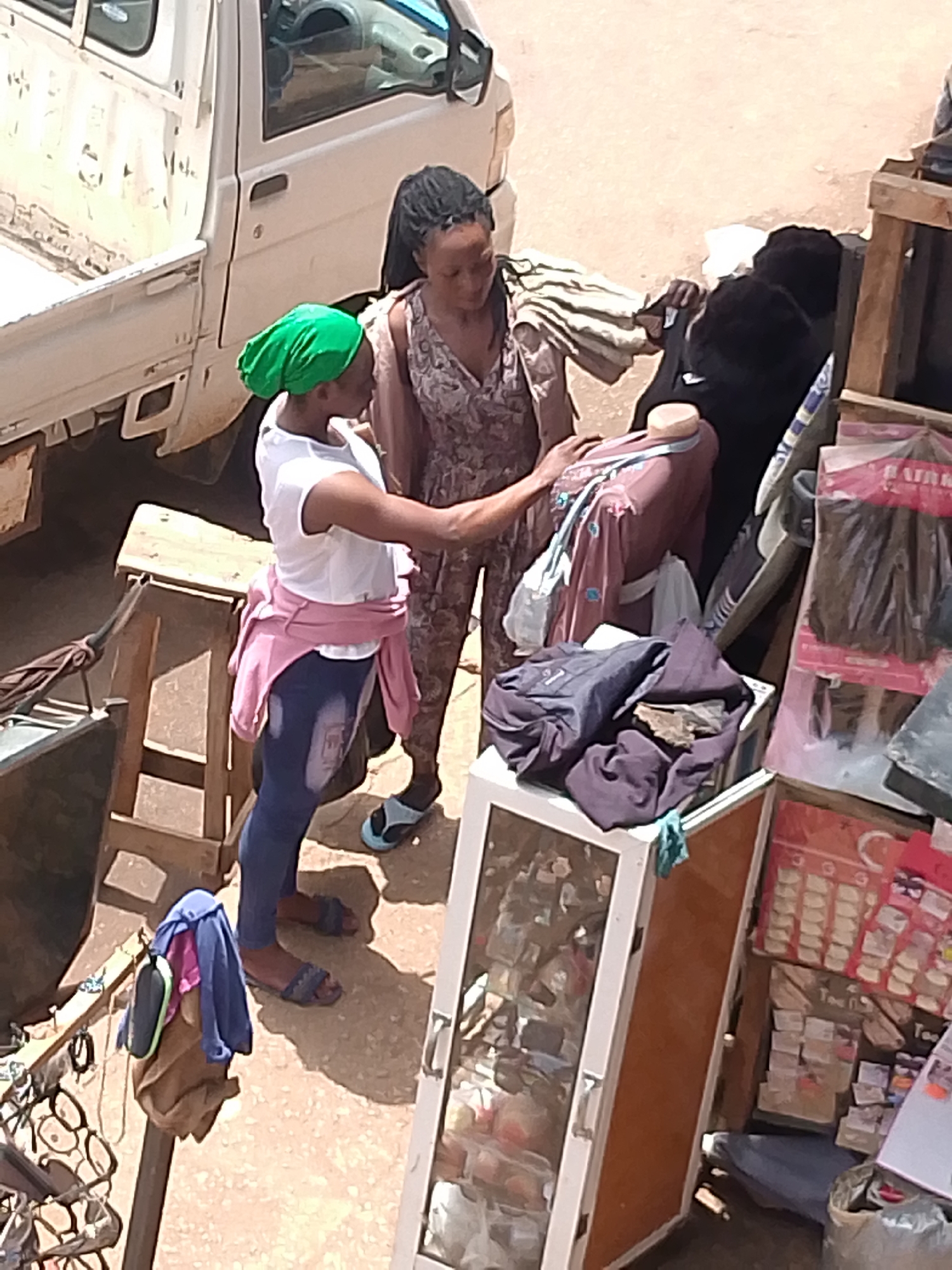 Two women stand next to a clothing display in an open-air market. One woman wears a green headscarf, a white t-shirt, blue jeans, and a pink sweater tied around her waist. She reaches toward a garment on a mannequin. The other woman wears a patterned dress. The display consists of clothing on a mannequin, folded garments on top of a white display case, and other clothing items. To the left is a white pickup truck. The foreground includes a display of eyeglasses. In the background are packaged hair extensions and other unidentified items. The location is identified as Bukuru, Nigeria.