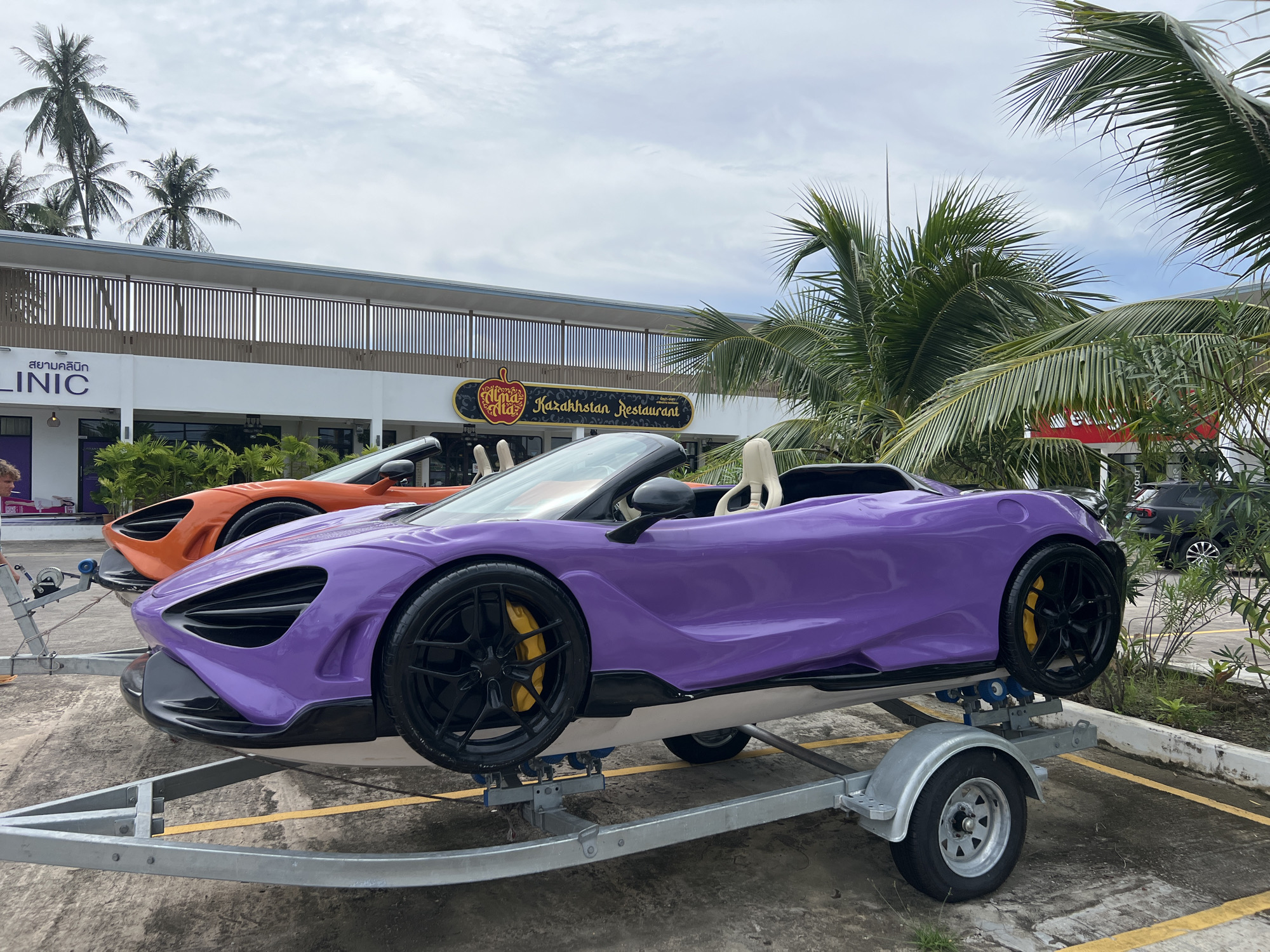 In Chalong, Thailand, two exotic sports cars, a purple McLaren and an orange McLaren, are positioned on trailers outdoors in front of a commercial building. The purple car is in the foreground, and the orange car is behind it. Both cars are convertible models. The building in the background features signage for "LINIC" and "Kazakhstan Restaurant." Palm trees and other vegetation are visible in the environment, and the sky is cloudy.