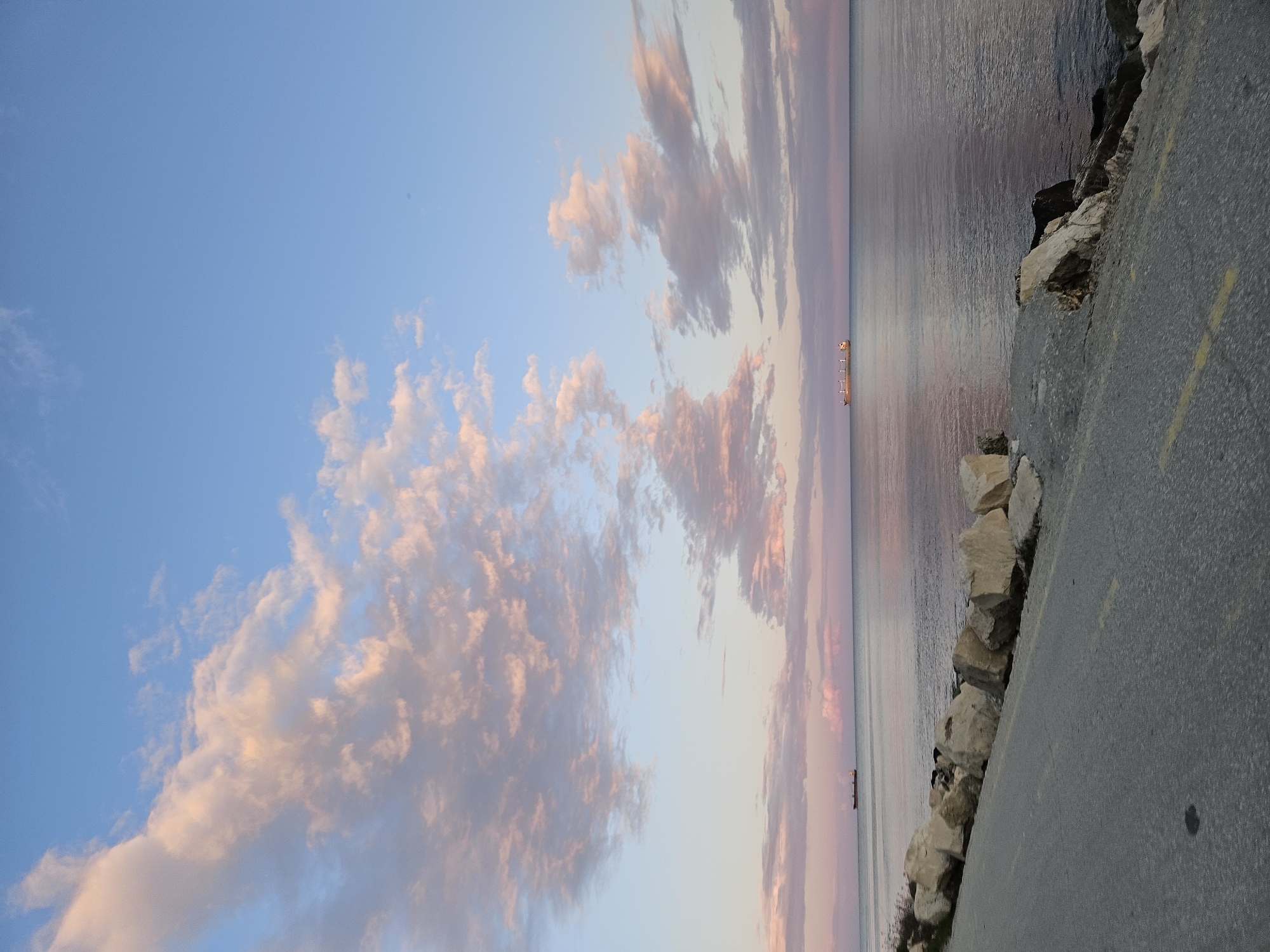 The image depicts a seascape under a sky with pastel-colored clouds, taken from a location in Varna, Bulgaria. The sky is a clear blue at the top, blending into shades of pink and orange near the horizon. The clouds are large and fluffy, illuminated with the same pastel hues. The sea is calm and reflects the colors of the sky. A long ship is visible in the distance on the water. Along the right side of the image is a paved walkway or road, with large rocks lining the edge where it meets the sea. A yellow line is visible on the pavement. The perspective suggests the photograph was taken from the ground, near the edge of the walkway.