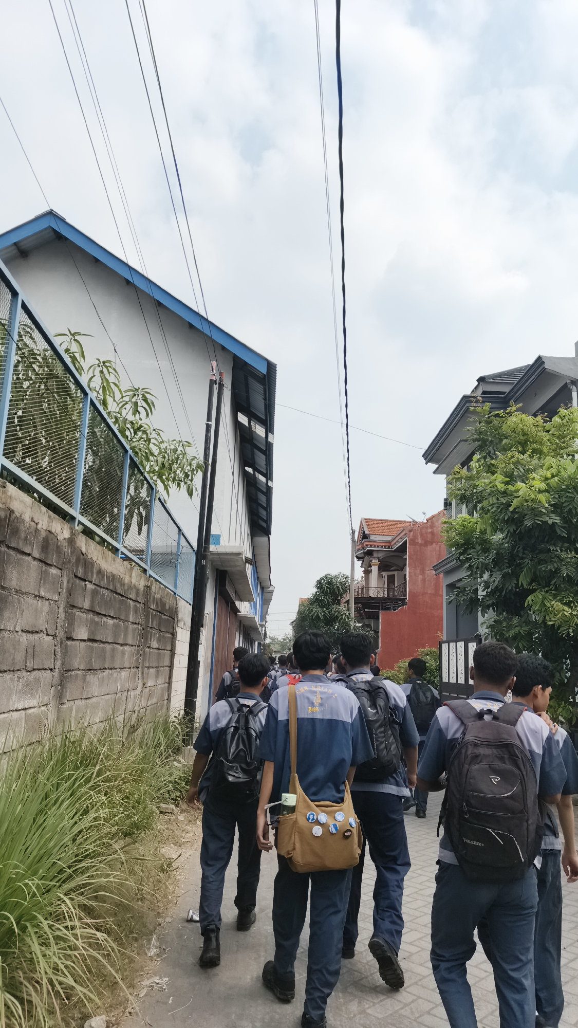 The image depicts a group of young people, presumably students, walking down a narrow, paved path between buildings. The path is bordered by a concrete wall on the left, with tall grass growing along its base. On the right side of the path, various residential buildings are visible, one with a distinctive orange-tiled roof and balconies. The students are all wearing similar blue and gray uniforms and carrying backpacks. Some have bags slung over their shoulders. Overhead, power lines stretch across the sky, which is partly cloudy. The scene appears to be in a semi-urban or suburban area. The context suggests this is a scene from Katrungan, Indonesia, as implied by the provided information, likely depicting students on their way to or from school.
