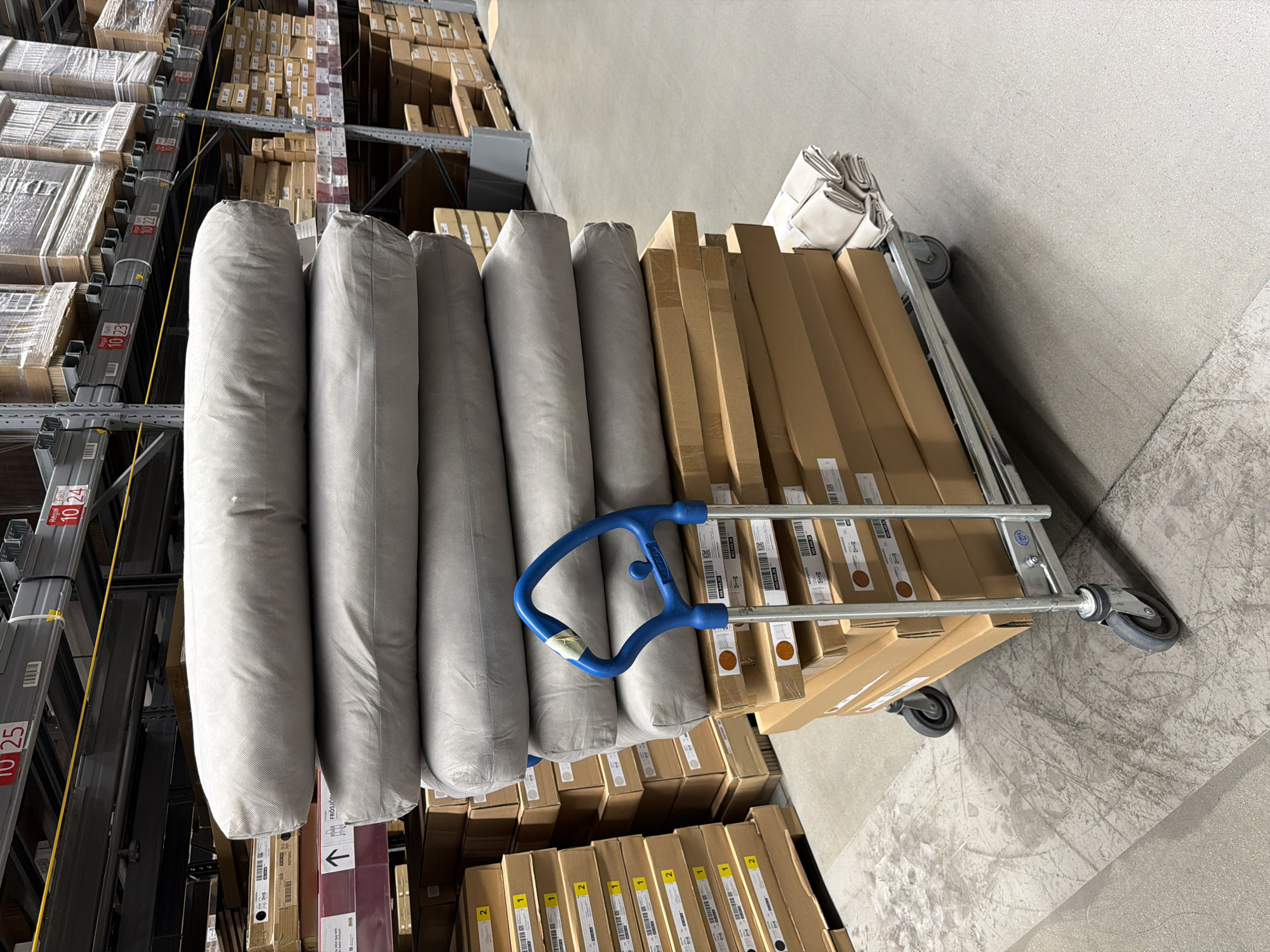 The image depicts the interior of a large warehouse, possibly a retail store, located in Kaiserslautern, Germany. Racks of stacked cardboard boxes extend into the background, while a flatbed trolley loaded with goods is in the foreground. The trolley is filled with gray cylindrical items, possibly packaged bedding, and stacks of long, rectangular cardboard boxes. The trolley has a blue handle and silver frame, with small wheels at the corners. The environment suggests a storage or distribution area, with a focus on inventory and logistics.