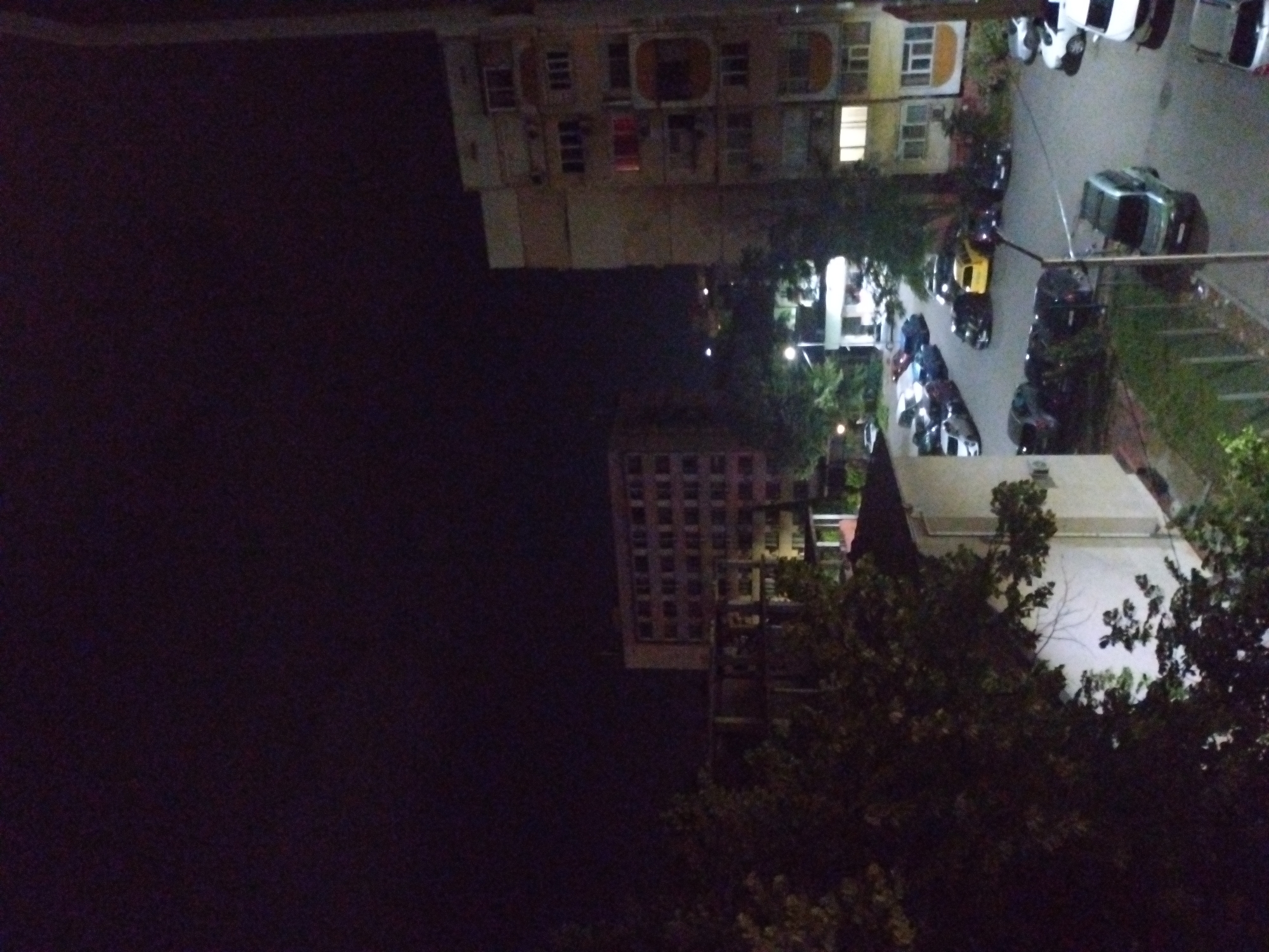 The image presents a nighttime urban scene in Stara Zagora, Bulgaria, captured from an elevated vantage point. The dark, cloudless sky occupies the upper portion of the frame. The environment is illuminated by artificial lighting, including streetlights and lights from buildings, casting a general yellowish-green glow on the ground and infrastructure.
In the mid-ground, a complex of asphalted roads and parking lots is visible, densely populated with parked vehicles. A bright yellow compact car is centrally positioned among other vehicles, including multiple dark-colored sedans and SUVs, and a silver SUV in the closer foreground. Several white cars are also present on the right side of the parking area.
The scene is bordered by several multi-story buildings. On the right, a prominent residential block with a light-colored facade features numerous windows and balconies, with lights visible in several apartments, indicating occupancy. A distinctive red light source is visible in an upper-floor window. Air conditioning units are attached to some balconies. In the mid-ground to the left, another multi-story building with a light facade and a grid of windows is partially obscured by dense foliage. A smaller, two-story white building with a dark roof is partially visible in the foreground-left, also partially hidden by trees.
Extensive green foliage from trees and bushes is scattered throughout the scene, particularly in the left foreground and between the buildings and parking areas. A streetlight pole stands centrally in the mid-foreground, contributing to the scene's illumination. No individuals are discernible within the frame.