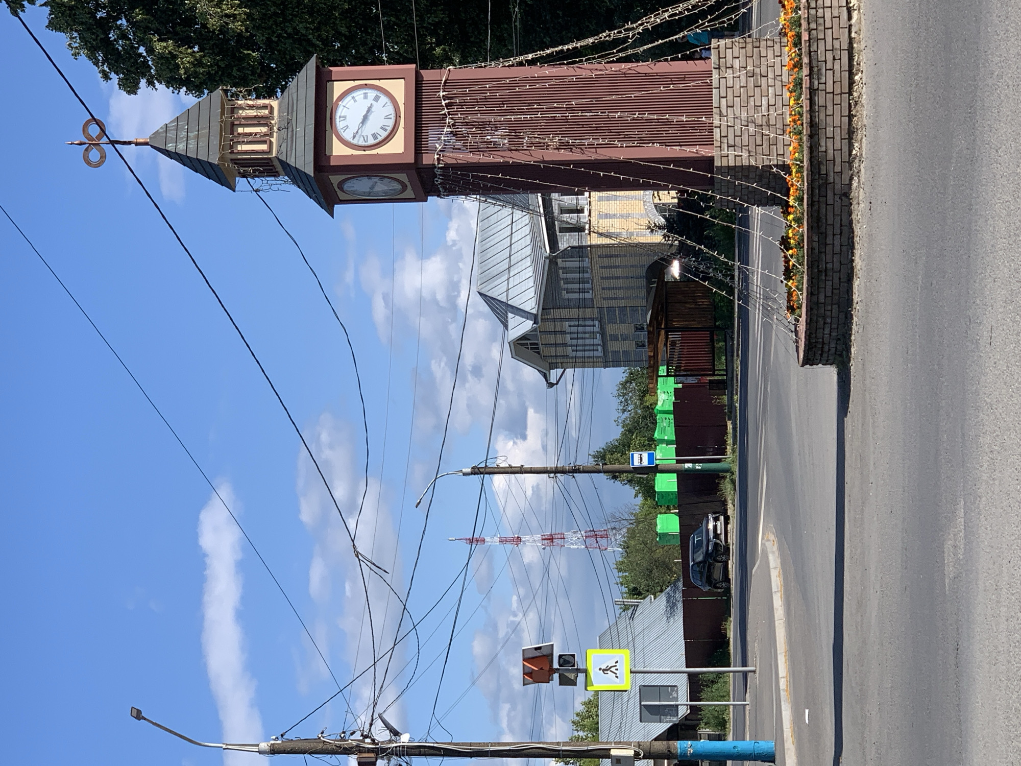 The image depicts an outdoor scene in a town, likely during the daytime under a clear sky with scattered clouds. The primary subject is a tall, reddish-brown clock tower with a peaked roof, adorned with decorative lights that appear to be turned on. The clock face is visible, showing Roman numerals and hands indicating a time. Next to the clock tower, a brick wall with a row of orange flowers runs along a paved road.
In the background, several buildings with distinctive architectural styles, including a yellow and grey building, are visible. A tall, red and white transmission tower stands in the distance. The scene is a mix of urban infrastructure and natural elements, with trees visible behind the buildings.
Several traffic signs and utility poles with numerous wires are also present, suggesting a busy street or intersection. One sign is a pedestrian crossing signal, another is a bus stop sign, and there are green rectangular signs on poles. A dark-colored car is parked on the side of the road. The overall impression is a functional, perhaps slightly aged, town setting on a bright day. There are no people or animals clearly visible in the image.
The location is identified as Pavlovo, Russia, which provides a geographical context for the scene.