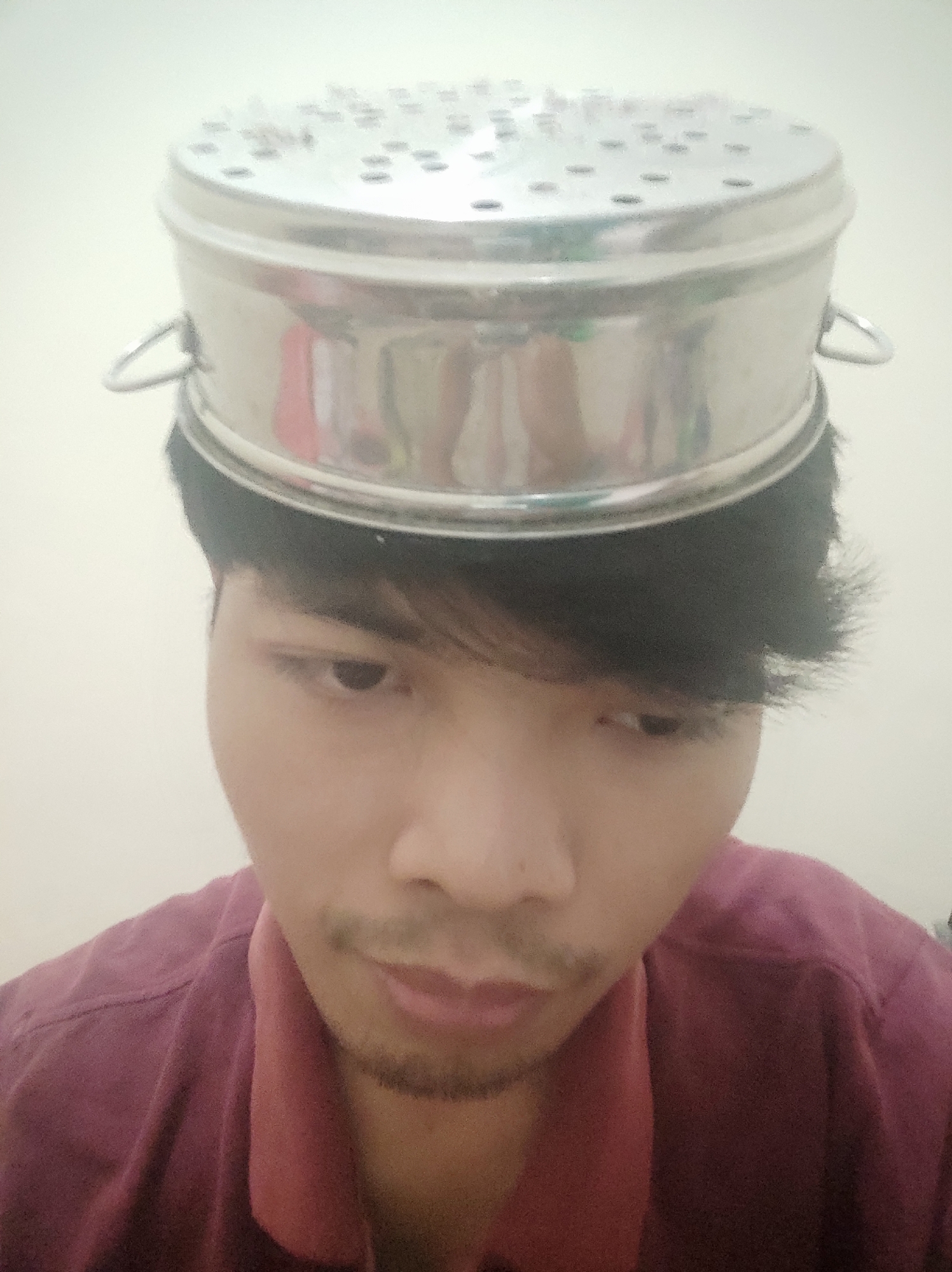 A person is wearing a stainless steel steamer basket as a hat. The person is looking down, with their face partially obscured by the basket. The background is a plain, light-colored wall.
