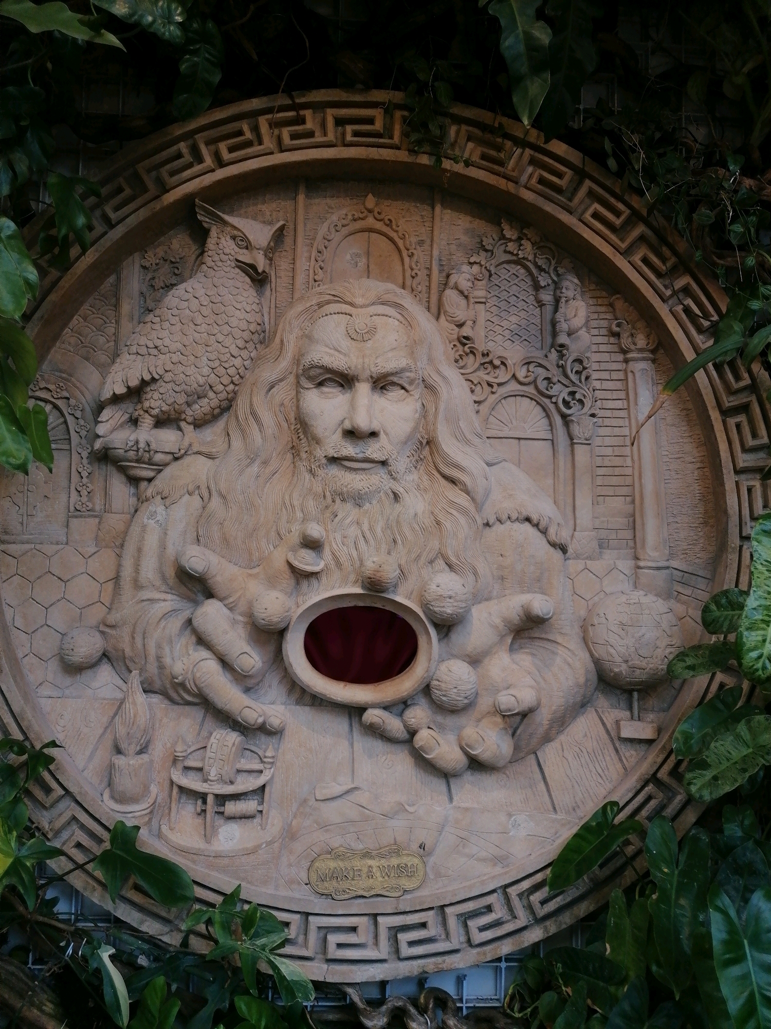 This is a medium shot of a bas-relief sculpture with a man depicted with a long beard and flowing hair, wearing a crown. He is holding a red orb in his cupped hands. To his left, an owl-like creature with detailed feathers perches on a column. To the right of the man, a globe with continents is depicted. Below the man, a candle, a cartographic instrument, and a plaque reading "MAKE A WISH" are carved. The sculpture is circular and framed by a Greek key pattern border. The artwork is set against a backdrop of lush green foliage, suggesting an outdoor or garden setting. The scene is dimly lit, with dappled light filtering through the leaves. The sculpture appears to be made of stone or concrete. The location is identified as Ubud District, Indonesia.