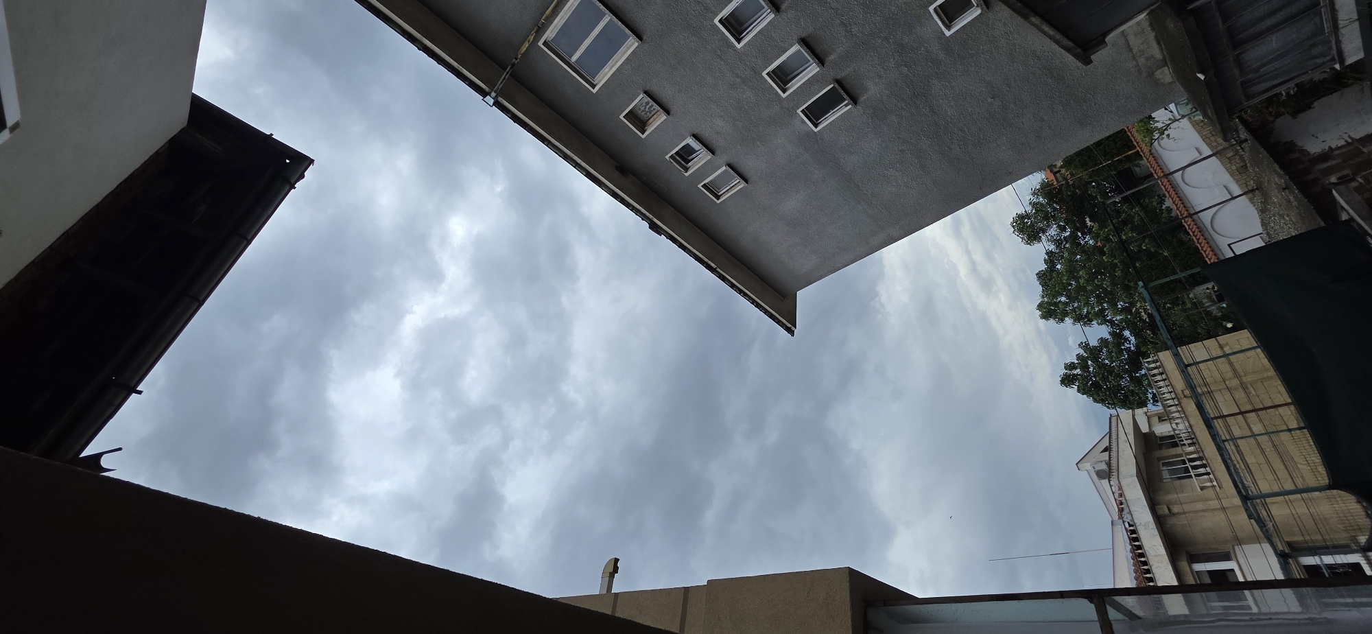 The image presents an upward-looking view of a cloudy sky, framed by architectural elements of surrounding buildings. The perspective is rotated, giving the impression that the buildings are angled diagonally against the sky.
In the upper-left and central portion, a grey-rendered building dominates, featuring multiple rectangular windows of various sizes, some appearing open, others closed. A dark gutter system is visible along its lower edge. To the immediate left, a section of a dark-colored roof or overhang partially borders the frame.
On the right side, further in the background, a lighter-toned building (possibly beige or light yellow) is visible, partially obscured by green trees and what appears to be a metal fence or trellis in the mid-ground. Another dark structure or awning is faintly visible in the immediate foreground on the lower right.
The sky is heavily overcast with dense, grey clouds, suggesting a cloudy day without direct sunlight. A tiny dark speck, possibly a bird or a distant drone, is discernible in the upper-right portion of the sky. A dark, curved fixture is present on the lower-left dark roof, and a vertical pipe or vent rises from the bottom edge towards the middle-right. The scene captures a quiet, urban landscape on an overcast day, consistent with a setting in Plovdiv, Bulgaria. No people are visible, and there is no discernible text.