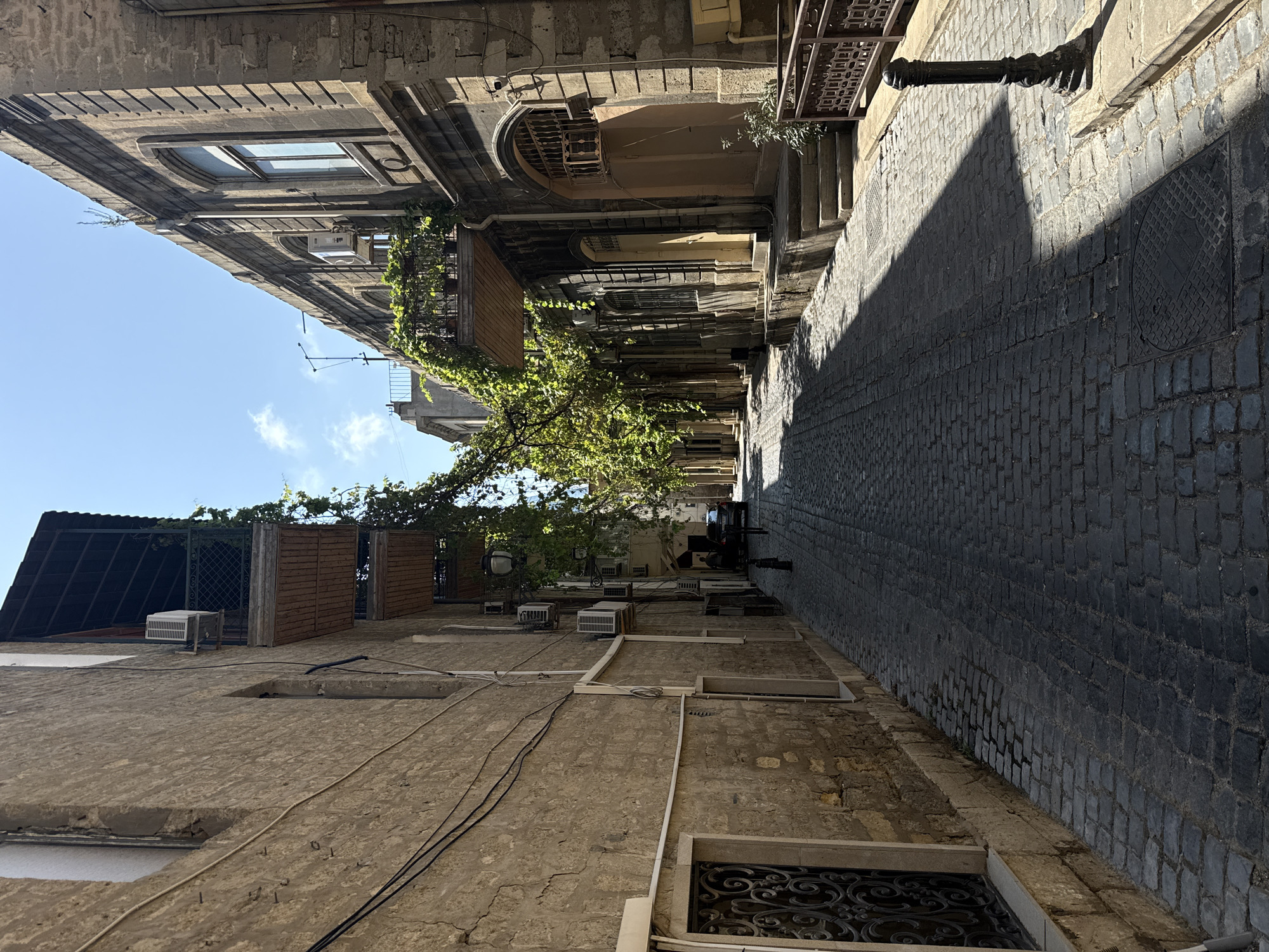 The image depicts a narrow, cobblestone street in the Sabail Raion of Azerbaijan. The street is lined with old, stone buildings, some of which have balconies overgrown with green vines. Several air conditioning units are visible on the facades of the buildings. In the distance, a car is parked on the street, and a person is walking away from the camera. The sun casts strong shadows across the cobblestones, indicating a bright, sunny day. A decorative black lamppost stands on the right side of the street, near a brown metal railing.