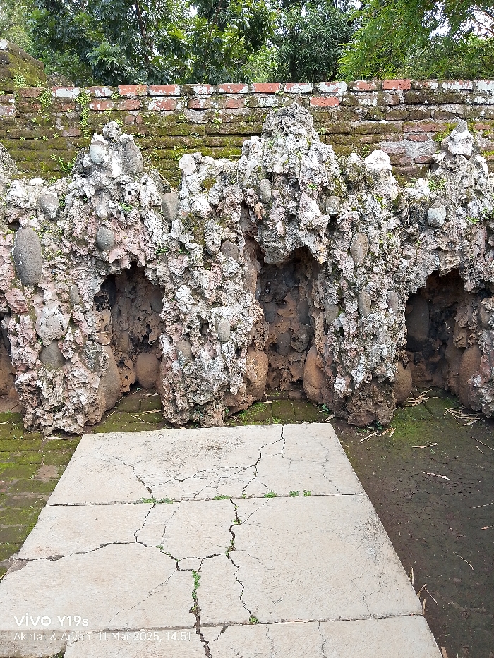 The image shows a stone wall with several openings, each resembling a small cave. The wall is built of irregularly shaped rocks, with some evidence of moss growth. The wall is constructed with red brick at the bottom and is located in Cirebon, Indonesia, at coordinates -6.737 latitude and 108.544 longitude. In the foreground is a cracked concrete floor. There are no people visible in the image. The scene appears to be a part of an ancient structure or temple complex. The wall is likely to be a part of a building in the city of Cirebon, Indonesia.