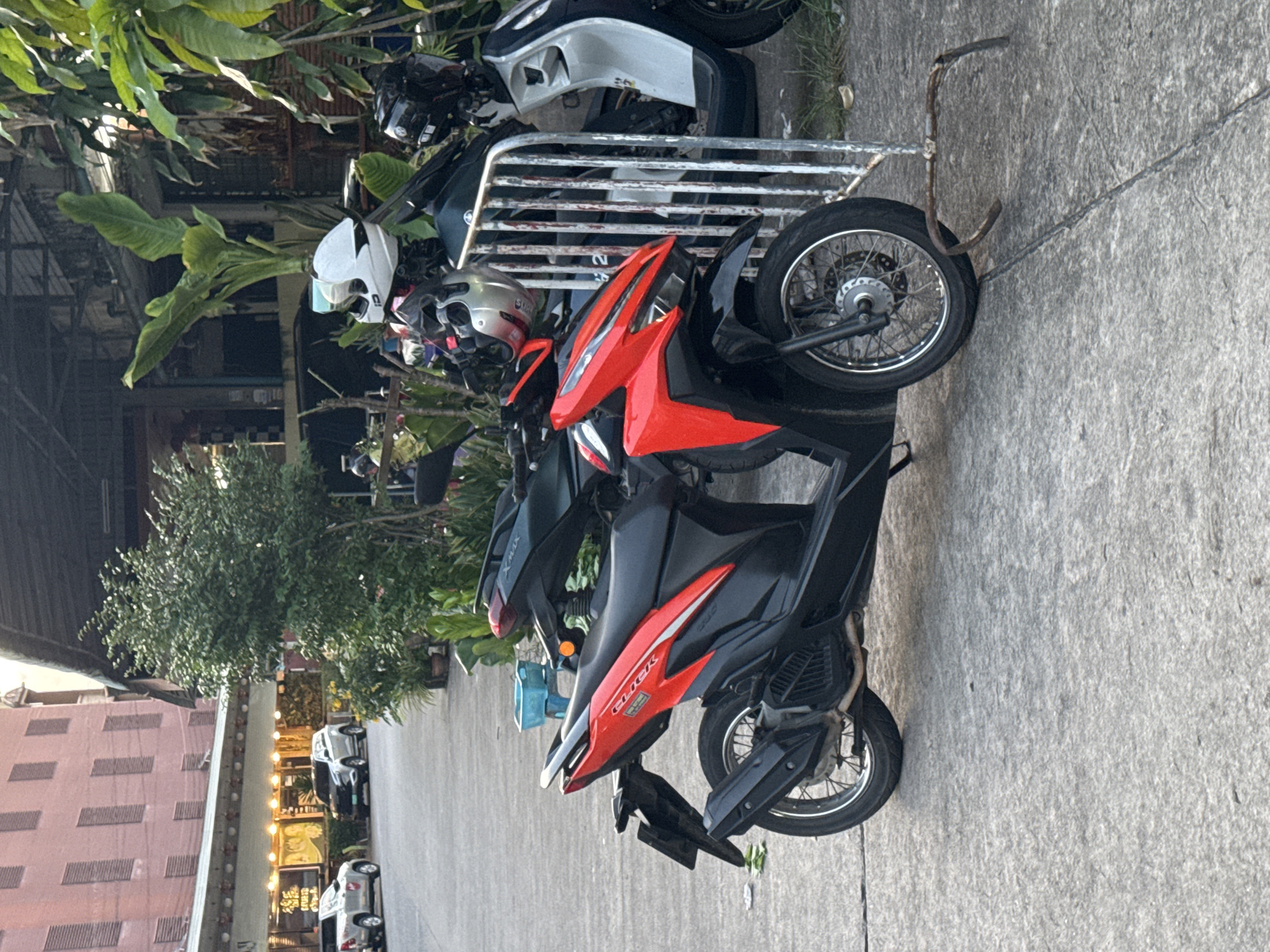 A red and black scooter is parked on a concrete street in front of a building with a covered walkway. Two other scooters, one dark green and one black and white, are parked behind it. There are several potted plants and trees around the area. In the background, a multi-story building with pinkish walls can be seen. Cars are parked on the street in front of shops. This scene is consistent with an urban street in Phuket City Municipality, Thailand, likely in a commercial area with shops and services.