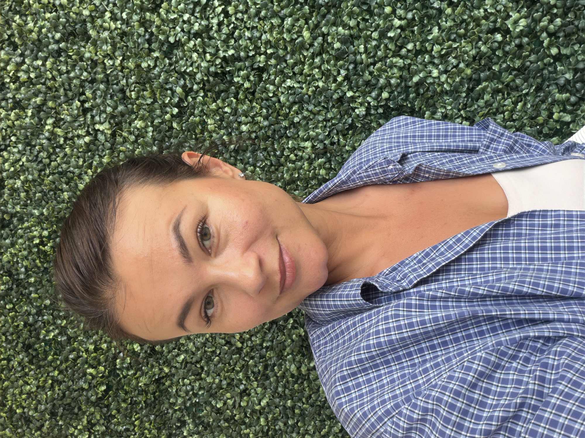 A woman with brown hair pulled back and wearing a blue and white plaid shirt over a white top smiles at the camera. She is standing in front of a green leafy wall, possibly an artificial hedge. The image is a selfie taken from a slightly upward angle. The background appears to be an outdoor setting, possibly a courtyard or a terrace.