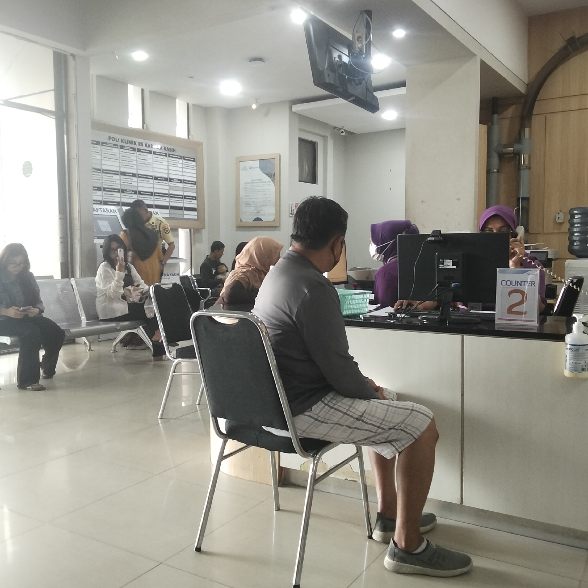 The image shows the interior of what appears to be a hospital or clinic waiting area. In the foreground, a man wearing a grey long-sleeved shirt and checkered shorts is seated on a metal chair, facing away from the camera. To his left, another metal chair is visible.
In the mid-ground, several people are seated on similar chairs. To the far left, a woman with glasses is looking at her phone. Next to her, a woman wearing glasses and a white cardigan is holding a phone up, possibly taking a picture or video. Behind them, another person is seated.
Further back, to the right of the man in the foreground, there is a counter area. Two women are behind the counter. One woman, wearing a purple hijab and a medical mask, is using a phone and looking at a computer screen. A sign on the counter reads "COUNTER 2". The other woman behind the counter is mostly obscured, but appears to be wearing a headscarf.
On the wall behind the counter, there is a sign with text in Indonesian that reads "POLI KLINIK RS KAR A KASIH" and "AFTARAN". There is also a digital display screen mounted on the ceiling. The floor is tiled, and the overall lighting suggests an indoor environment, possibly with natural light coming from windows on the left side of the image.
Based on the text on the sign, "RS KASIH", this location is likely the "RS Kasih" hospital. The presence of "POLI KLINIK" indicates a polyclinic or outpatient department within the hospital. Without further context or external signage visible in the image, it is not possible to definitively pinpoint the specific city or location within Sukabumi, Indonesia, however, "RS Kasih" is a known hospital name in Sukabumi, Indonesia.