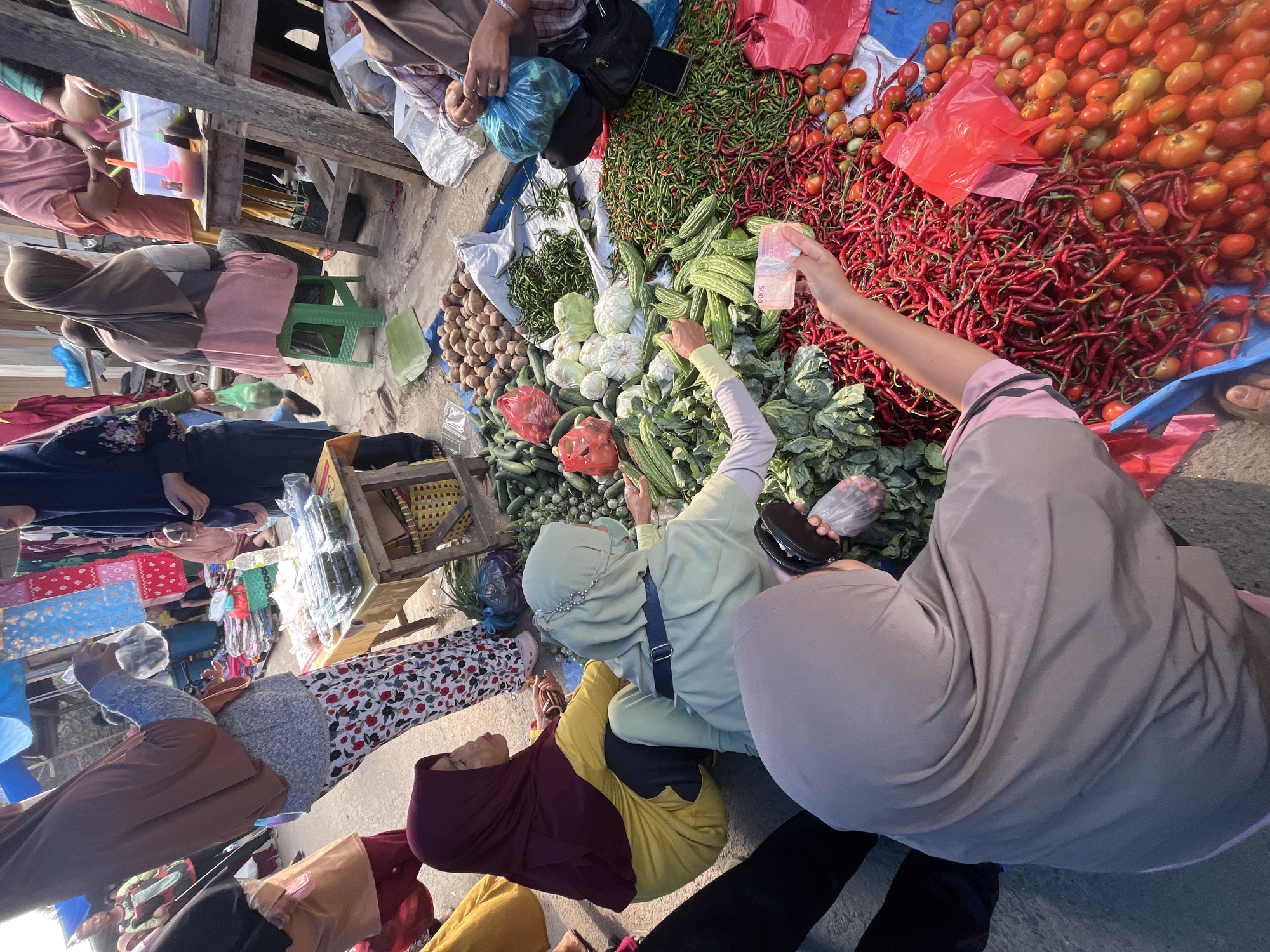 In Kemuning, Indonesia, a busy outdoor market scene unfolds, bathed in natural light. A group of women, predominantly wearing hijabs, are engaged in commerce. The focus is on a stall laden with fresh produce, including a large pile of red chilies, tomatoes, bitter melon, cabbage, cucumbers, and potatoes. One woman in a light green hijab, adorned with a beaded headband, is selecting vegetables, her hand reaching towards the produce. Another woman, wearing a burgundy hijab and yellow top, is also present, her gaze directed towards the activity. In the foreground, a woman in a grey hijab is holding out a 5000 Indonesian Rupiah banknote, seemingly in the process of a transaction. The market environment is evident with other stalls and shoppers visible in the background, suggesting a typical day of local trade.